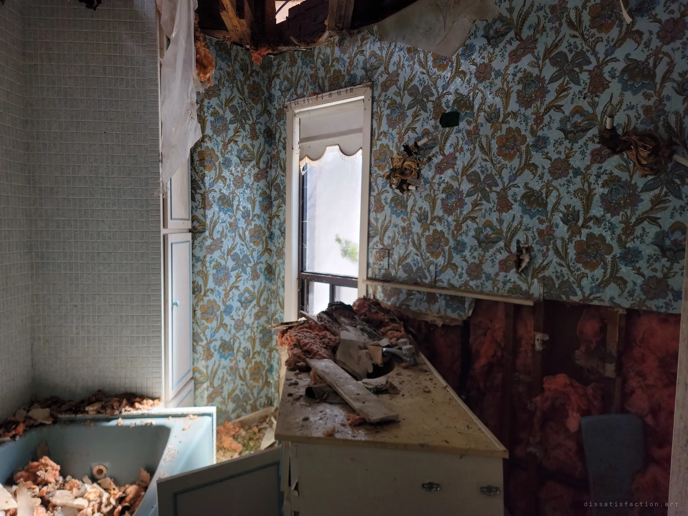 A bathroom with blue floral wallpaper and a gutted wall of pink insulation, the tub and vanity buried under fallen ceiling debris beside a narrow window.