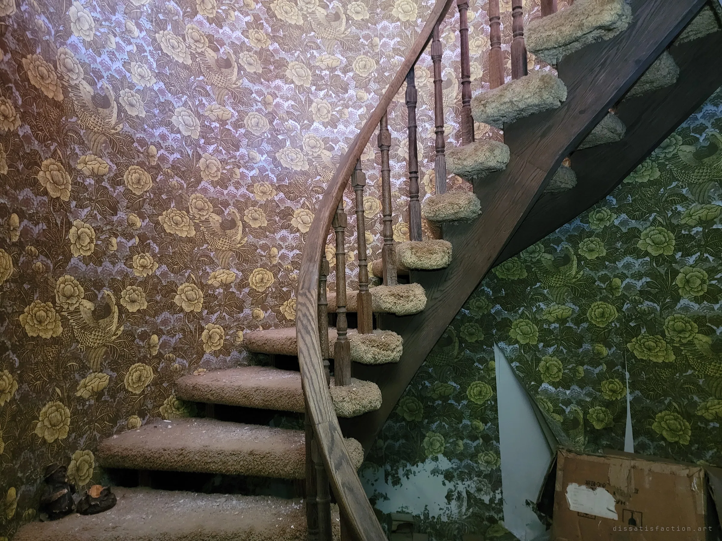 The foot of the curving wooden staircase, its shaggy carpeted treads climbing past walls of gold and green floral wallpaper peeling at the seams. An old cardboard box sits in the corner beneath it.