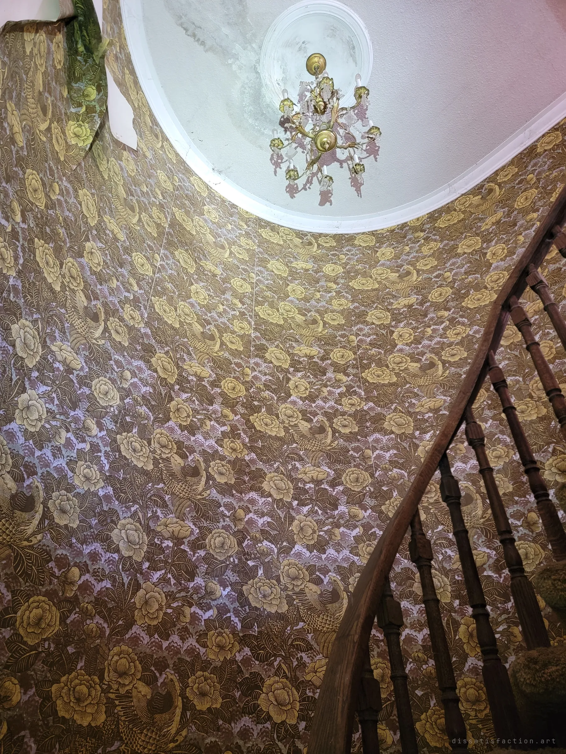 Looking up the curved stairwell to a small brass and crystal chandelier hanging from a water-stained ceiling medallion. Floral wallpaper sweeps around the whole shaft.