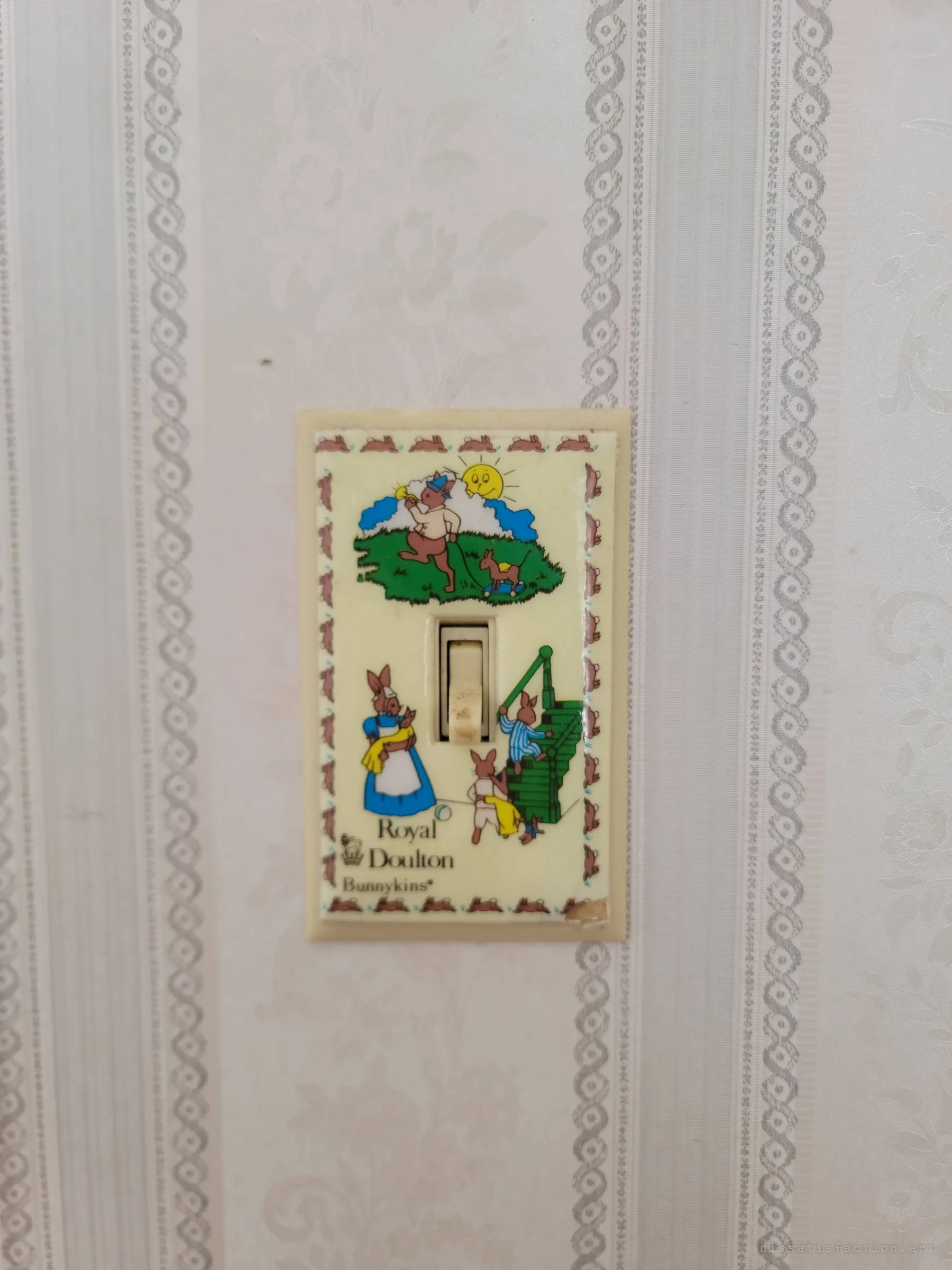 A child's light-switch plate printed with cartoon rabbits at play, set on pale striped damask wallpaper. A small trace of whoever grew up here.