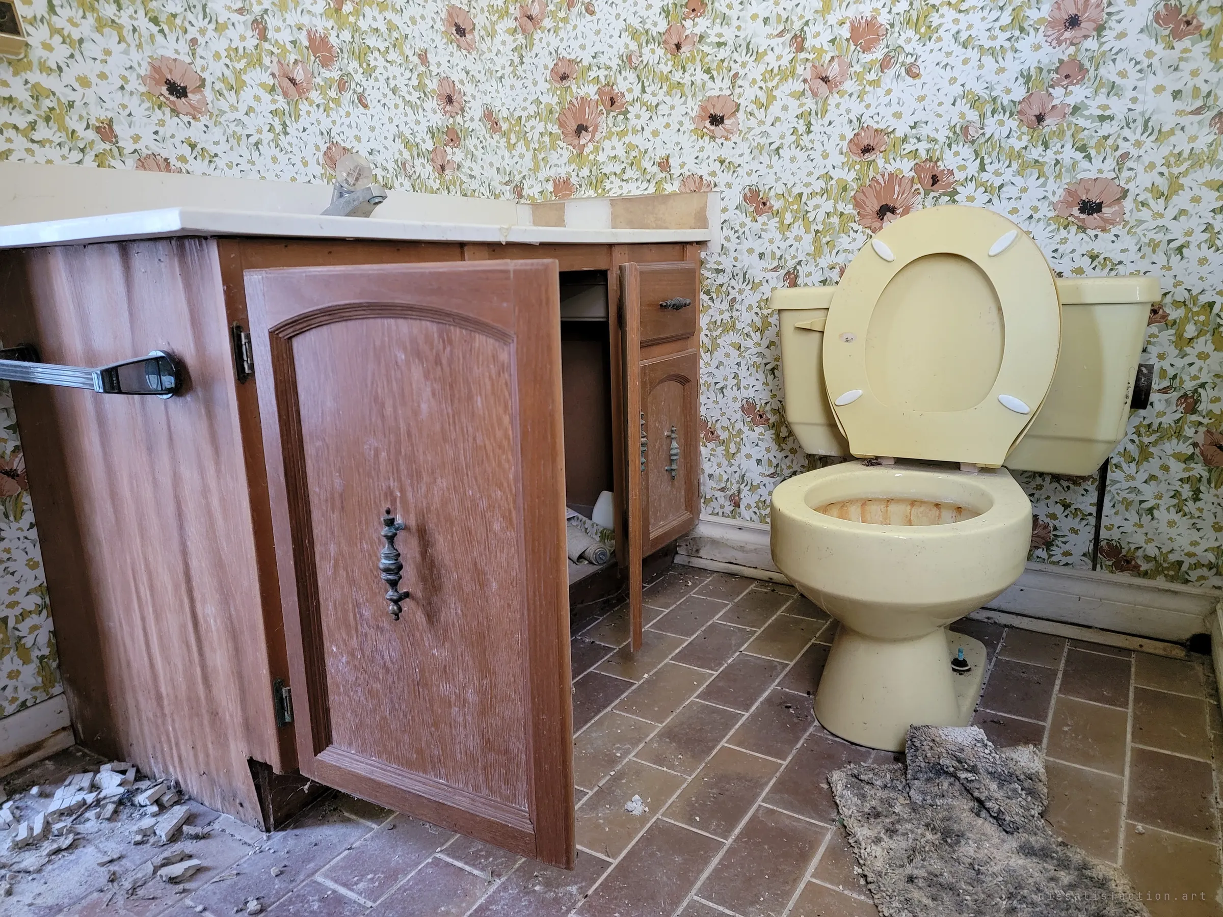 A bathroom with poppy-print wallpaper, a pale yellow toilet with its lid up and a wooden vanity with doors hanging open. A matted rug lies on the brick-pattern tile floor.
