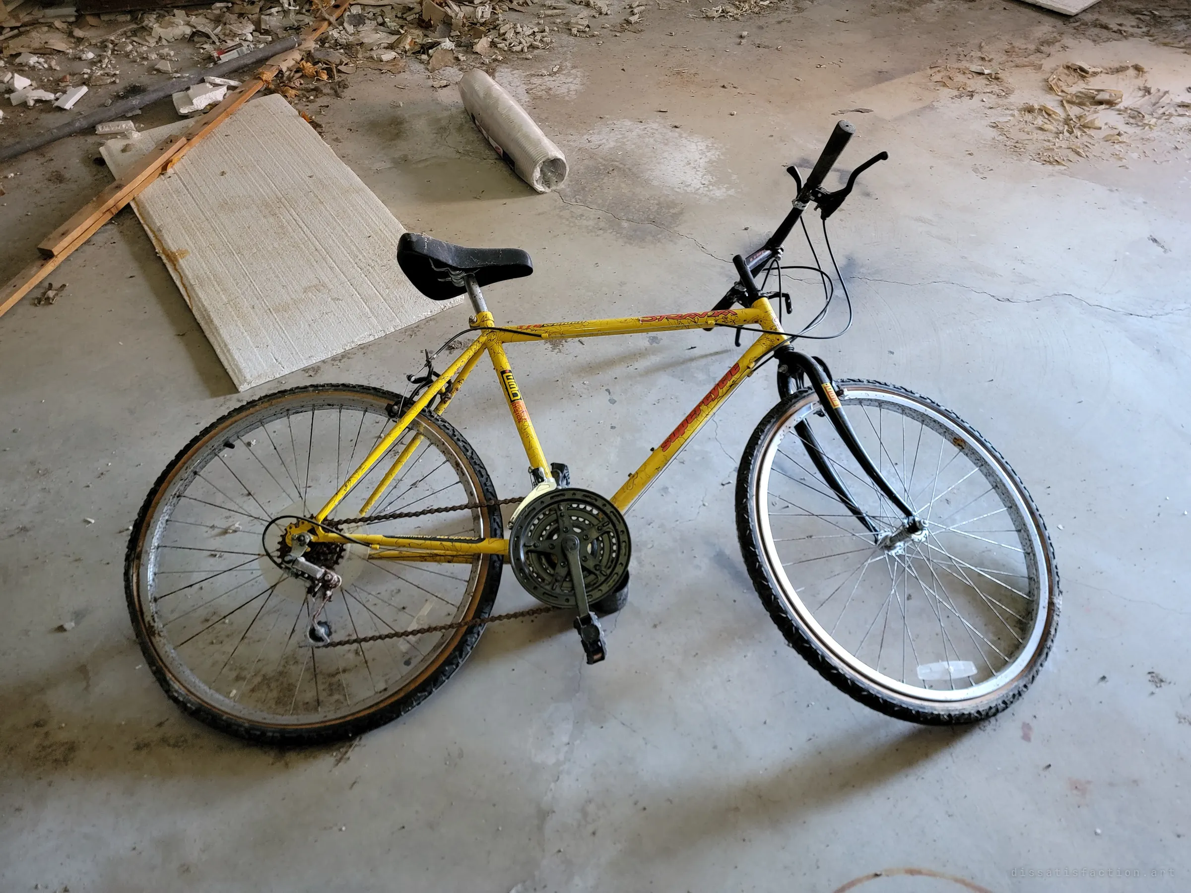A yellow mountain bike lying on its side on a cracked concrete floor, with foam board and debris behind it. Left where it fell.