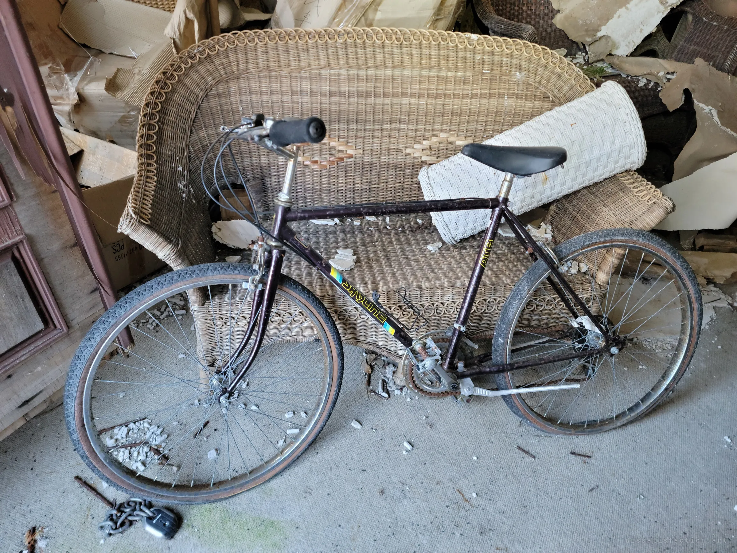 An old dark-framed bicycle leaning against a wicker loveseat, with a chain lock on the floor and torn cardboard piled all around. Storage slowly collapsing in on itself.