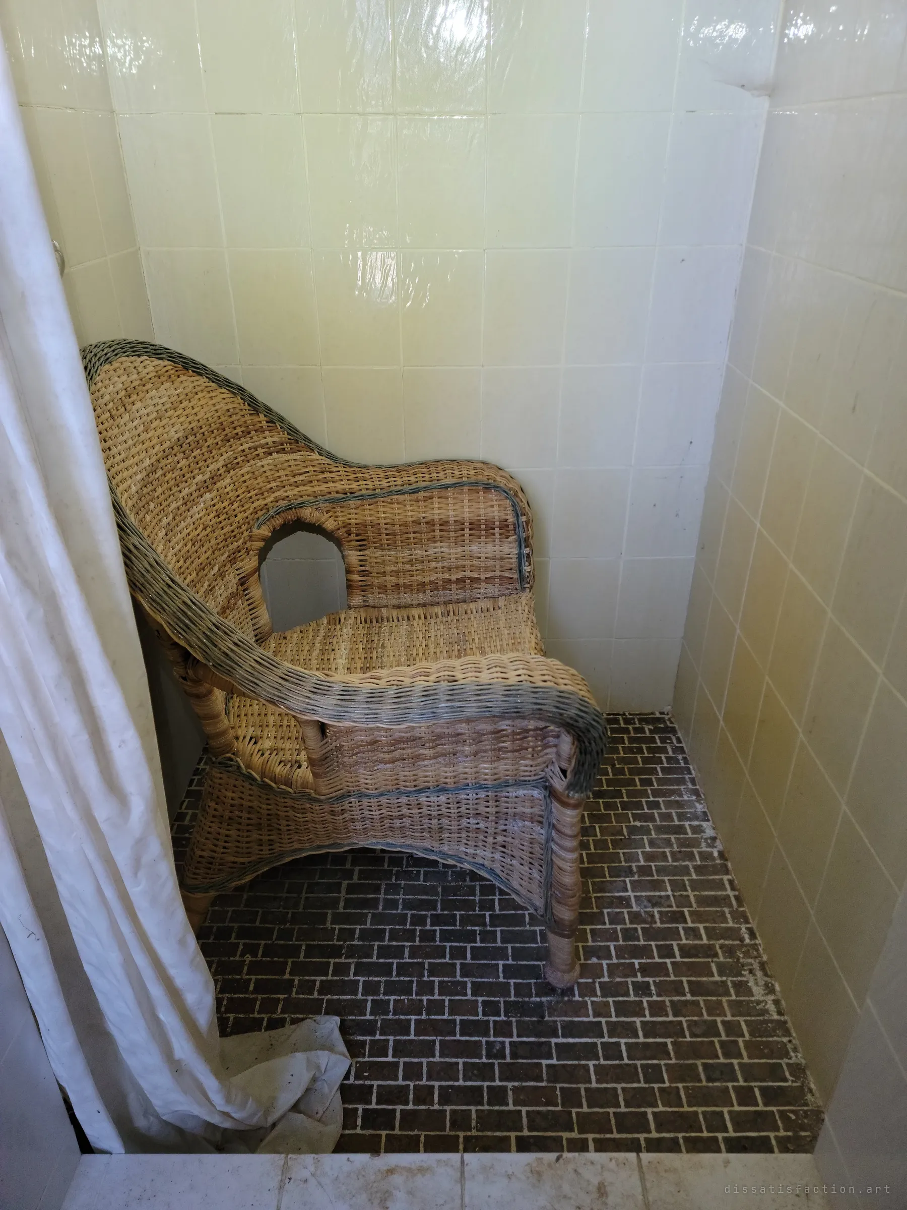 A wicker armchair sitting alone inside a tiled shower stall, the curtain pulled half aside. An empty seat in an odd place.