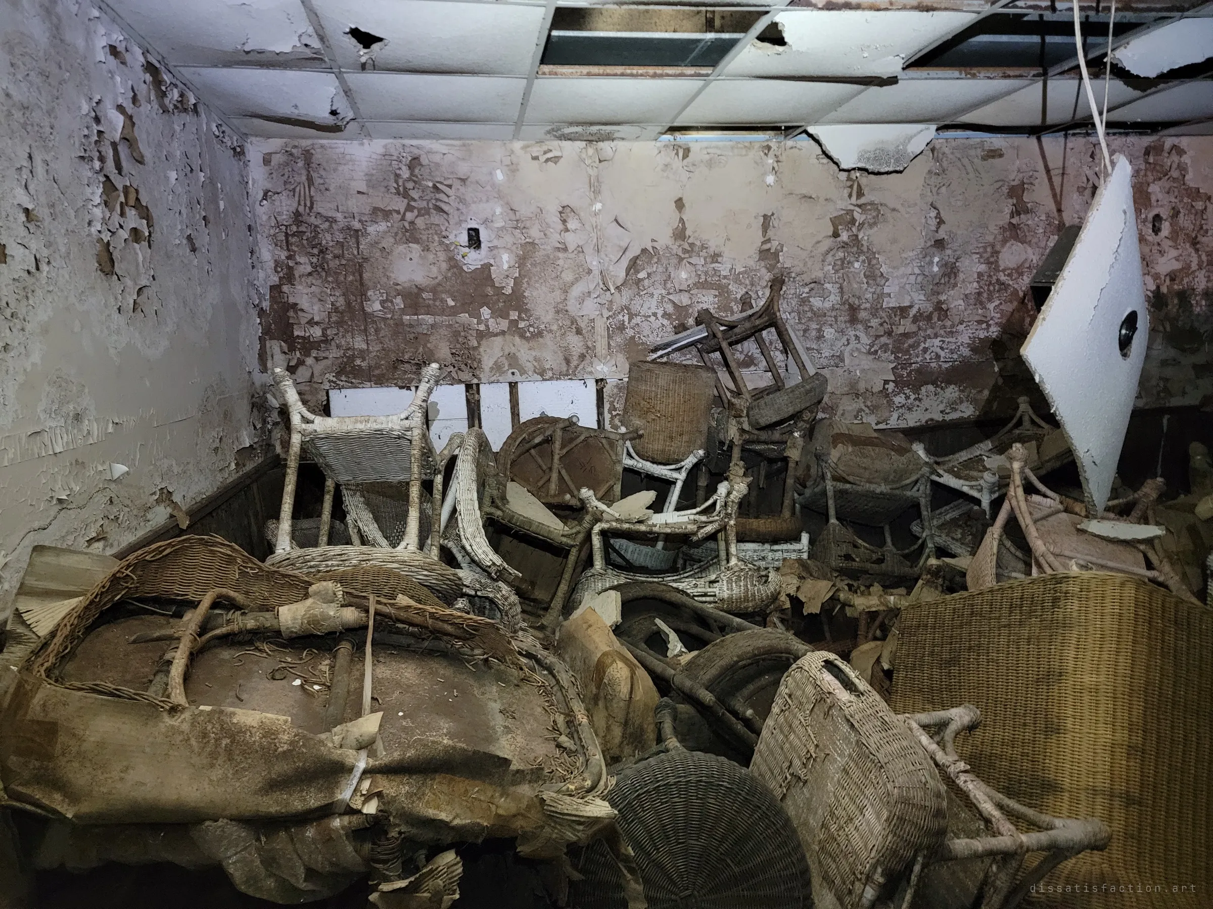 A low room crammed with heaps of broken wicker chairs under a collapsing drop ceiling, paint peeling off every wall. A fallen ceiling panel hangs at an angle on the right.