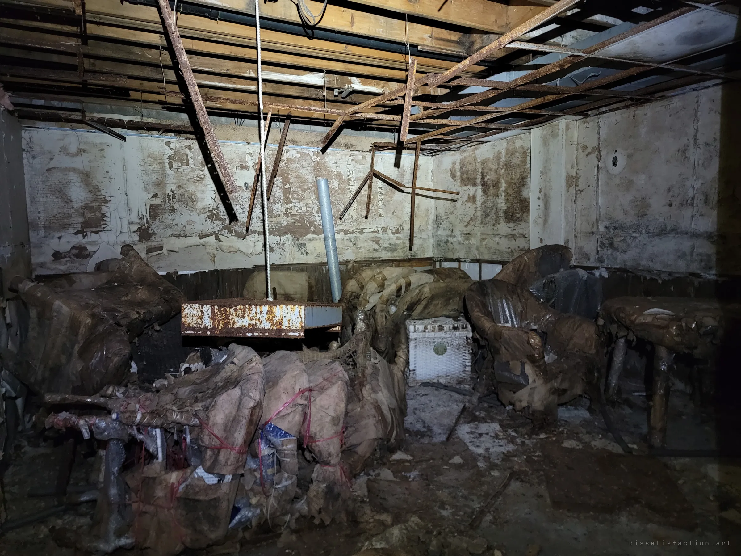 A flashlit basement room with its ceiling grid rusted and sagging over armchairs wrapped in rotting paper and twine. The wrapped furniture looks stored and forgotten, slowly melting into the damp.