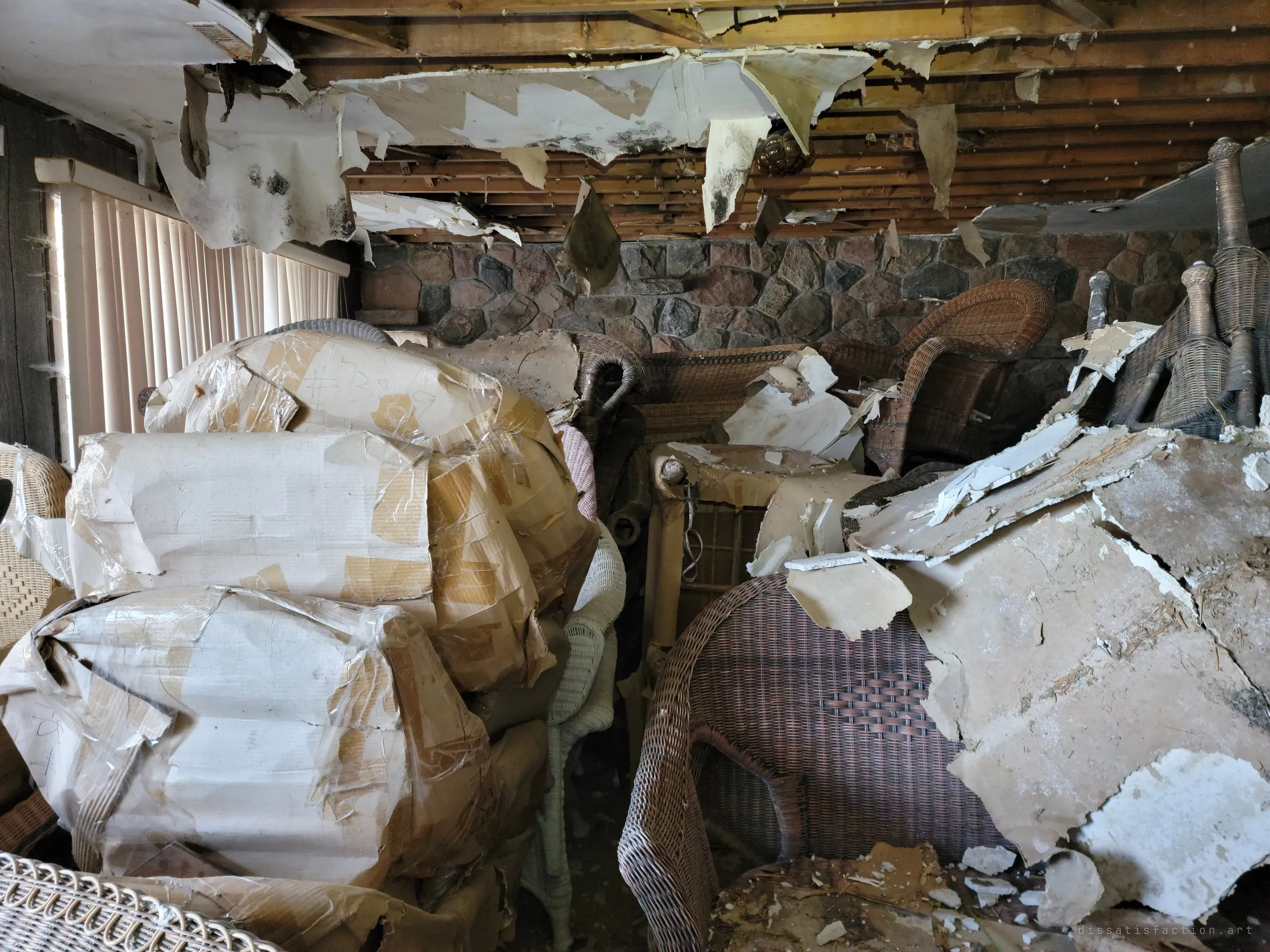 Wicker chairs and paper-wrapped bundles jammed against a fieldstone wall while ceiling drywall peels down in heavy sheets. Daylight through closed vertical blinds shows how packed the room is.