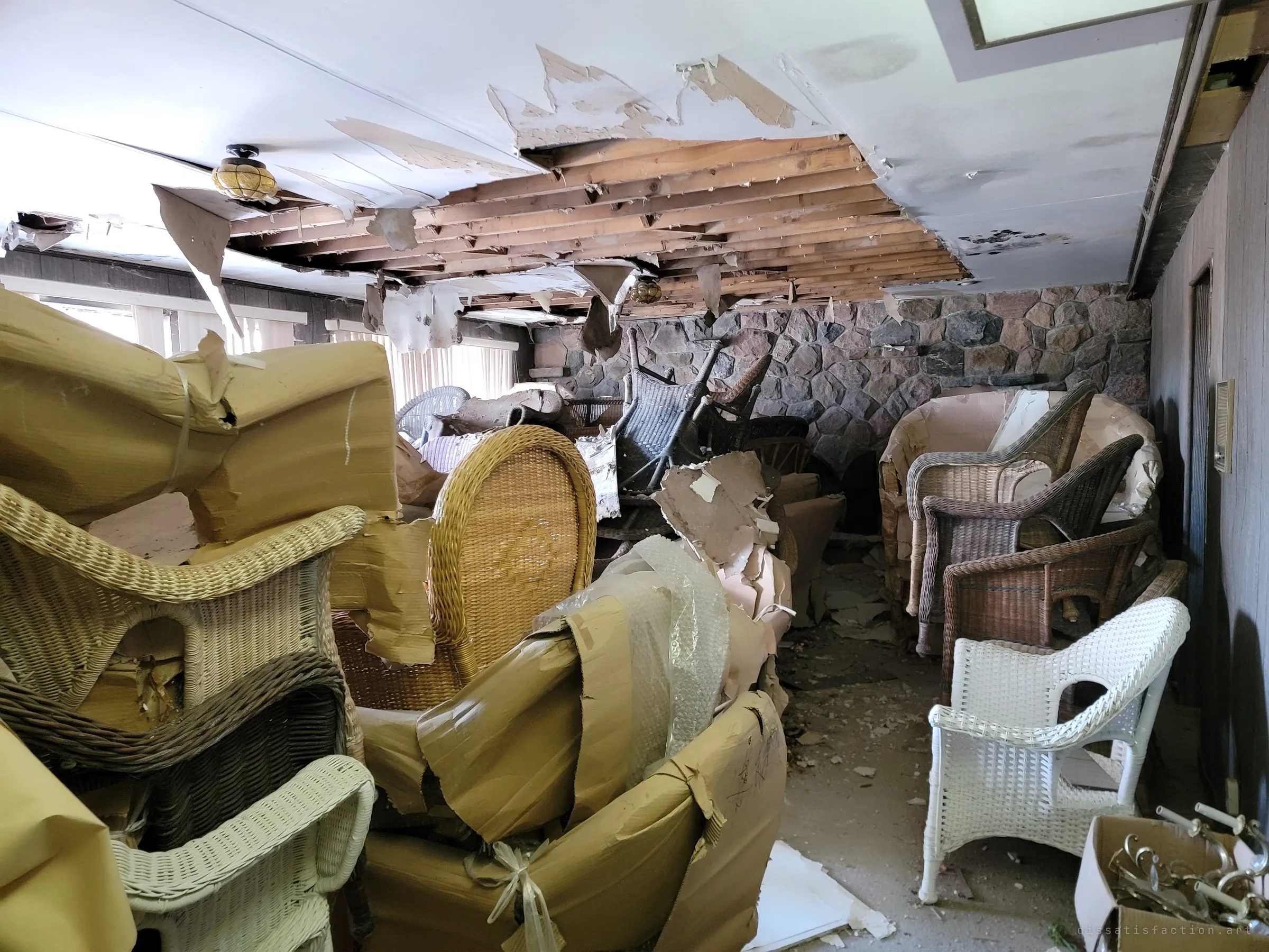 A long sunroom crowded with stacked wicker chairs under a torn-open ceiling, joists showing through. A fieldstone wall and closed blinds frame rows of furniture that never left storage.