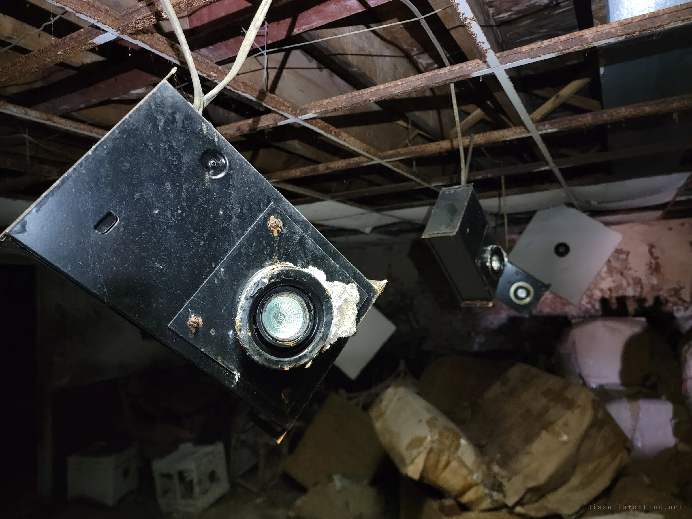Close-up of a black spotlight box dangling on its wires from a stripped ceiling, more fixtures hanging askew behind it. The lights that once lit a showroom now point at heaps of wrapped furniture.