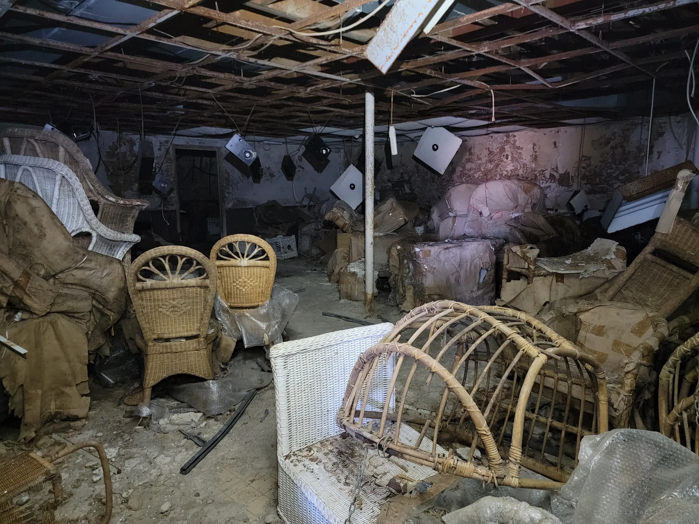 A low basement showroom full of wicker chairs and wrapped armchairs, square light fixtures hanging loose from a bare ceiling grid. A single white post holds up the room among the clutter.