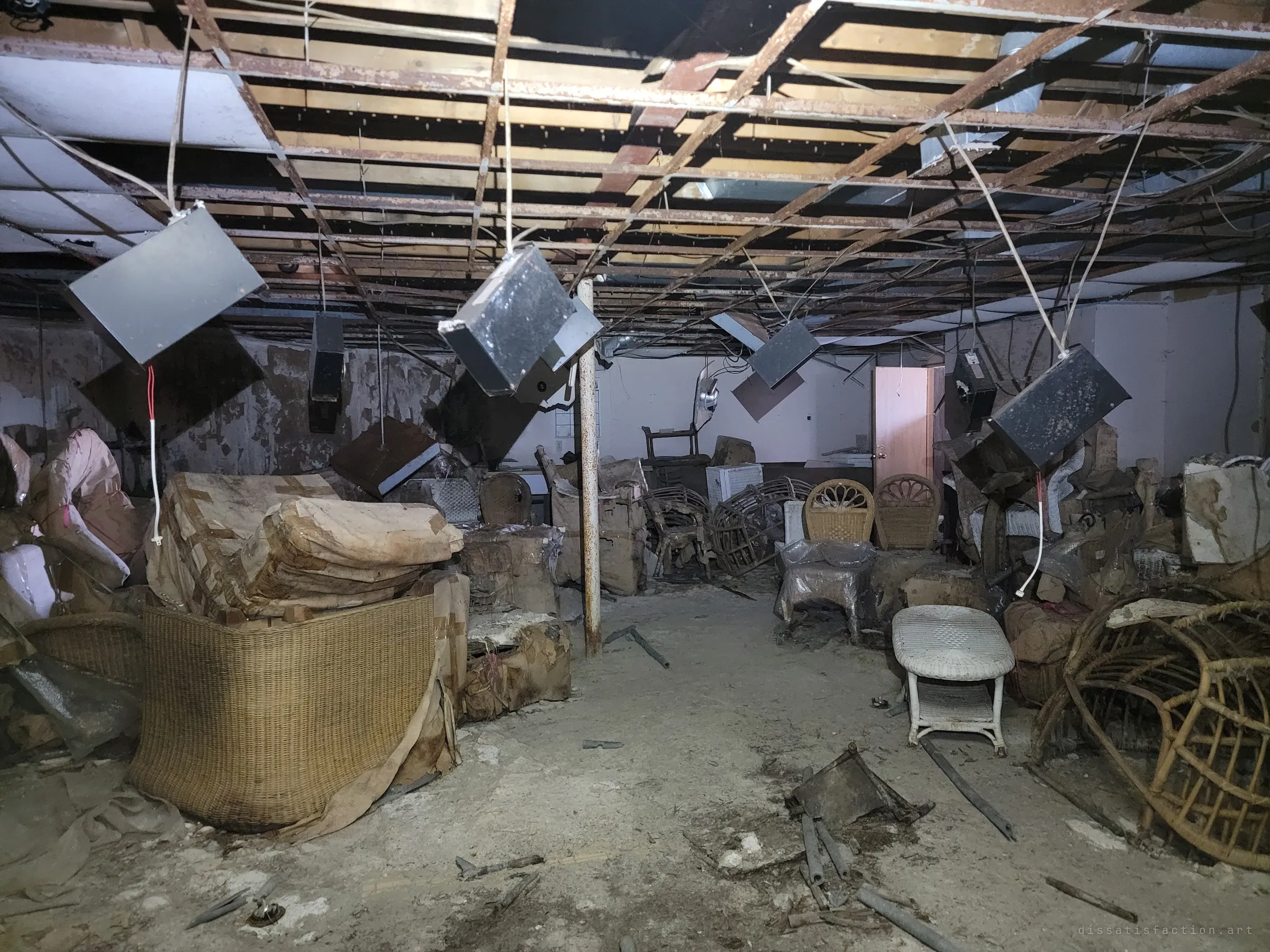 The same basement seen down its length, light boxes swinging on cables above a dusty aisle between piles of wicker and wrapped furniture. A pale door stands open at the back.
