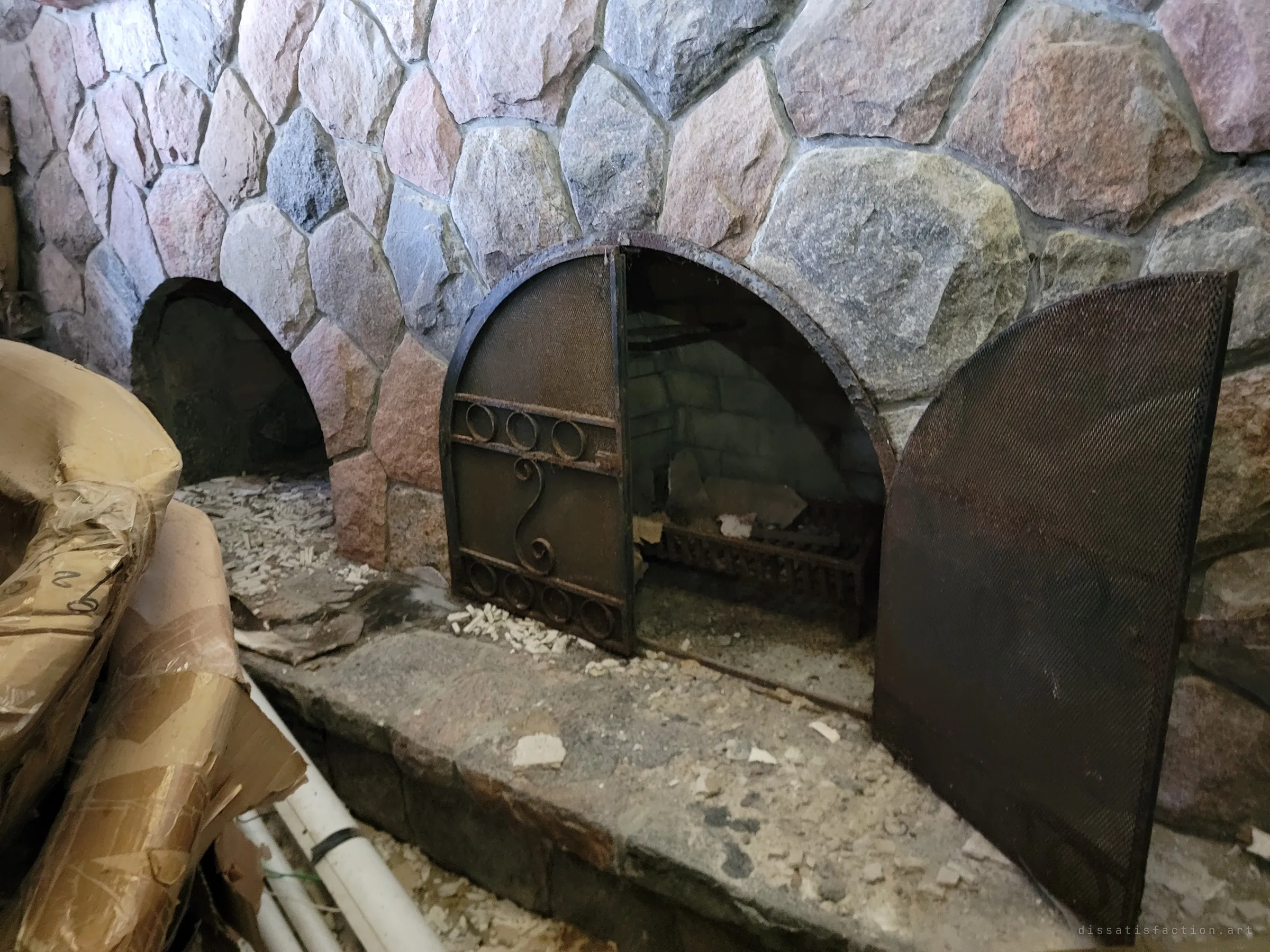 A fieldstone fireplace with two arched openings, one half-covered by a scrolled iron screen. Debris and wrapped furniture crowd the hearth that once warmed the house.