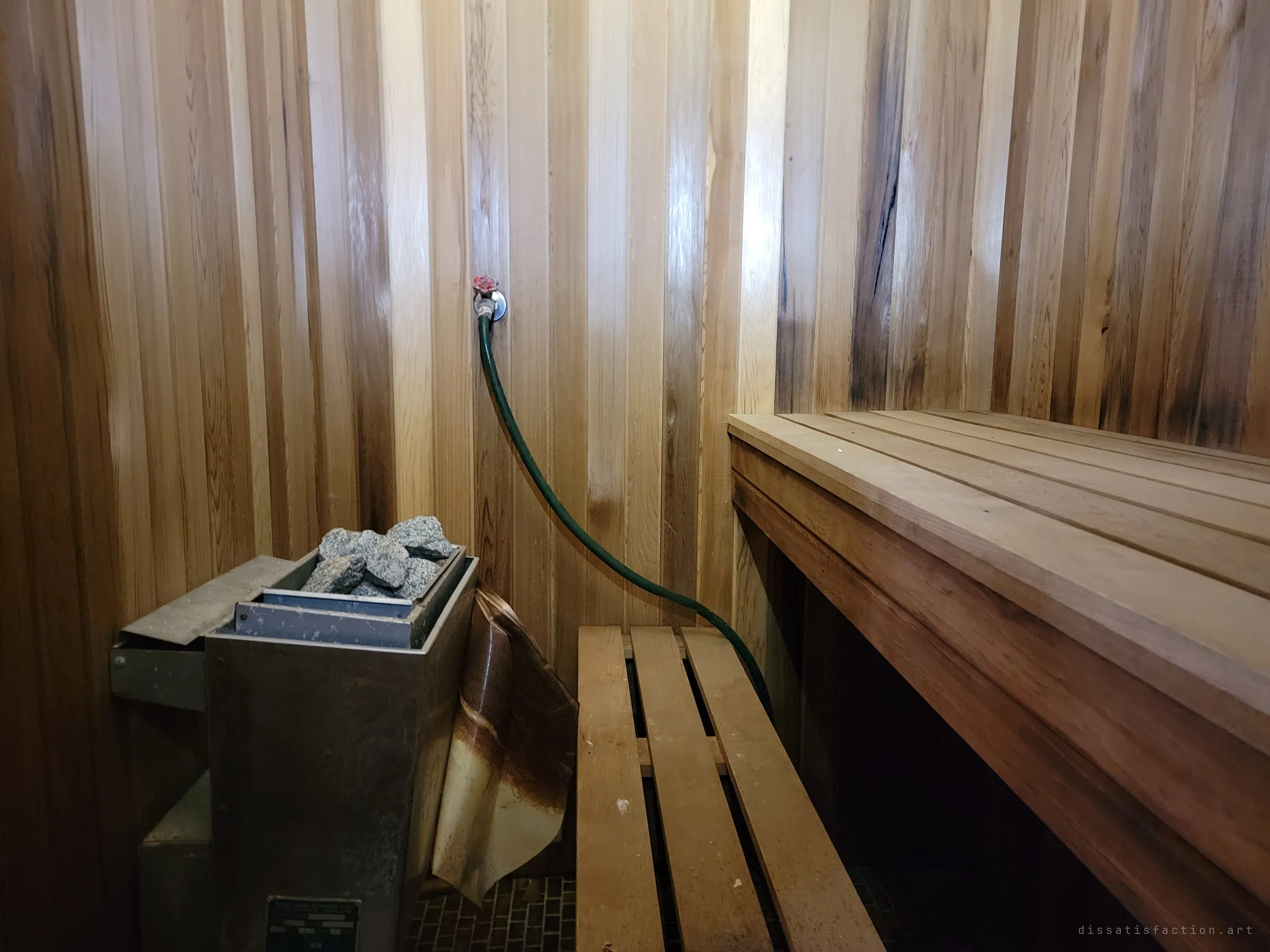 A cedar-panelled sauna with a stone-topped heater, slatted benches and a garden hose on the wall tap. It is strangely intact compared with the rot elsewhere in the building.