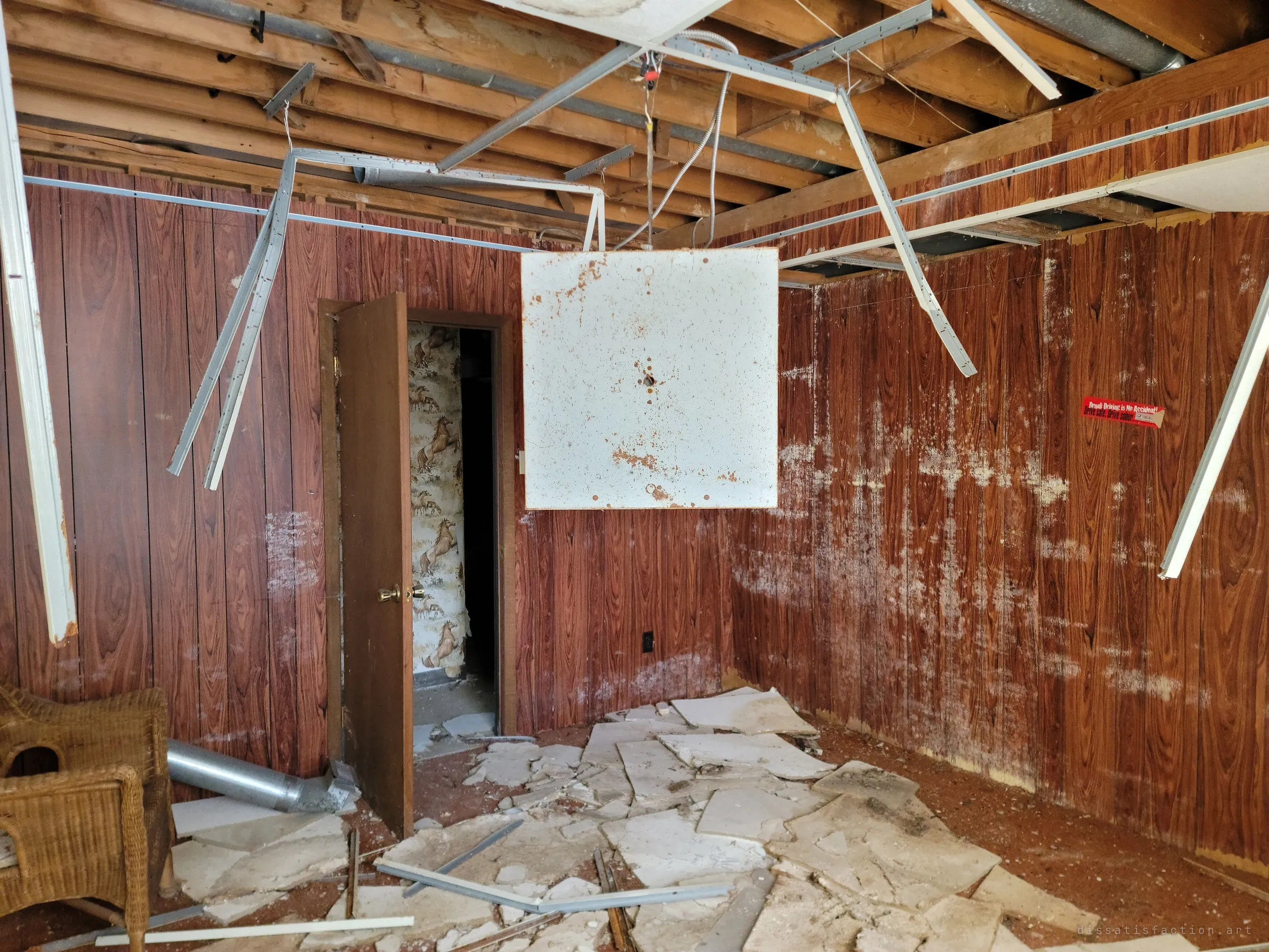 A wood-panelled room with ceiling tiles smashed across the floor and the metal grid twisted overhead. Through the open door, horse-patterned wallpaper hints at the property's past.