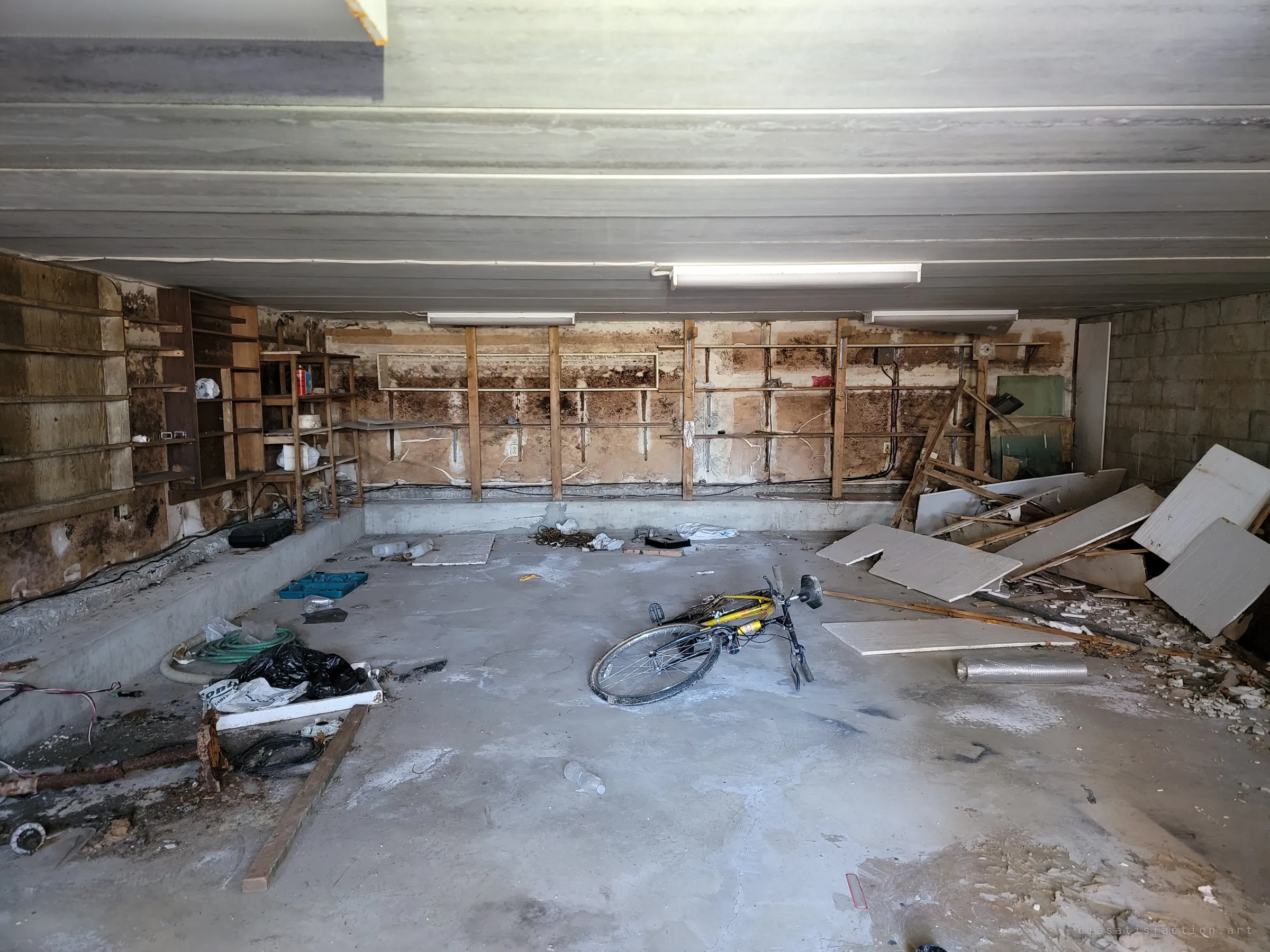 An empty garage with mould-blackened walls, bare shelving and a yellow bicycle lying on the concrete floor. Broken panels are heaped in the corner where someone gave up clearing it.