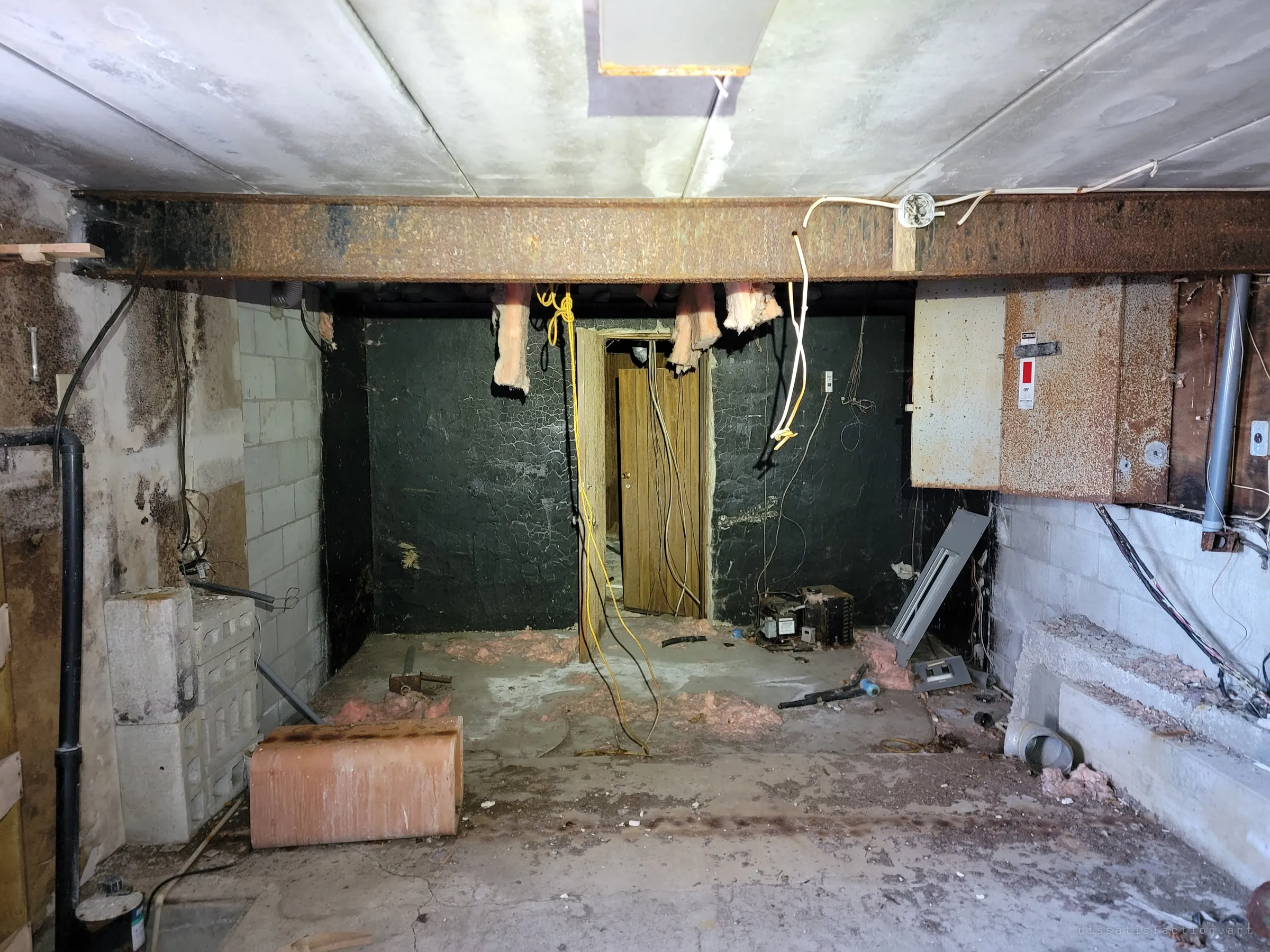 A cinder-block utility space under a rusted steel beam, torn insulation and loose wiring hanging toward a doorway in a black-painted wall. A rusted electrical panel sits switched off on the right.