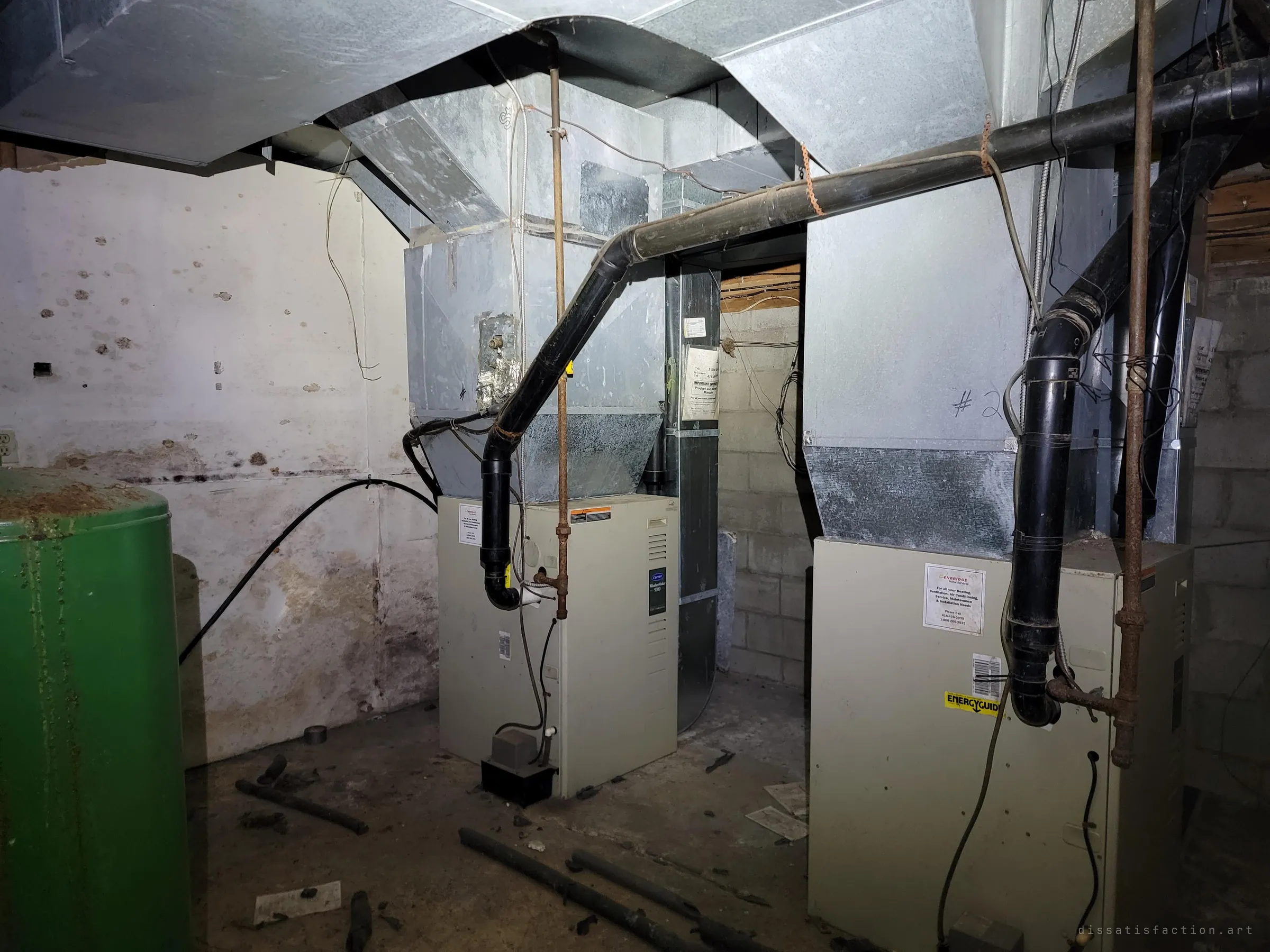 Two furnaces in a dim cinder-block utility room, tied into a tangle of galvanised ducts and black vent pipe beside a green tank. The machinery that heated the building sits cold and dusty.