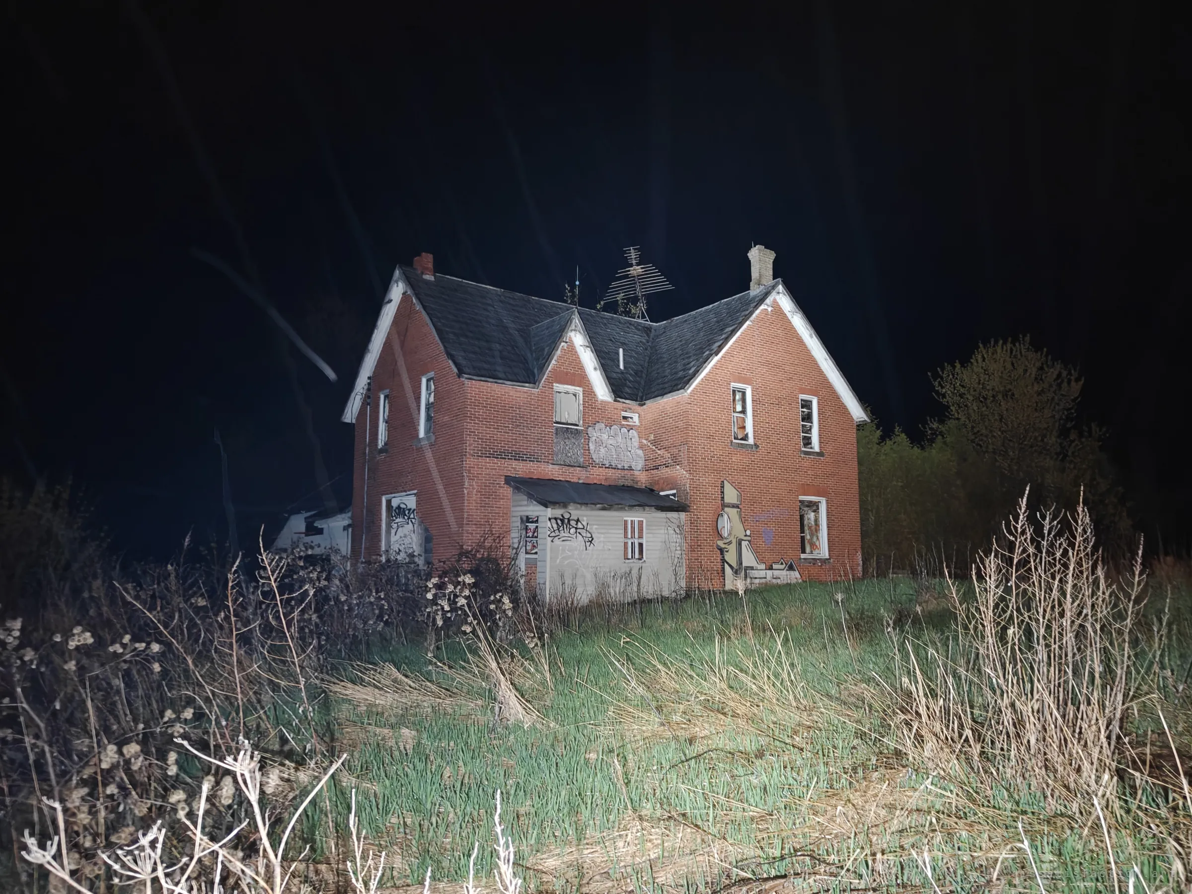 A two-gabled red-brick farmhouse lit up at night, its windows broken and walls tagged, standing in overgrown grass. The opening shot sets the scale of the empty house before going inside.