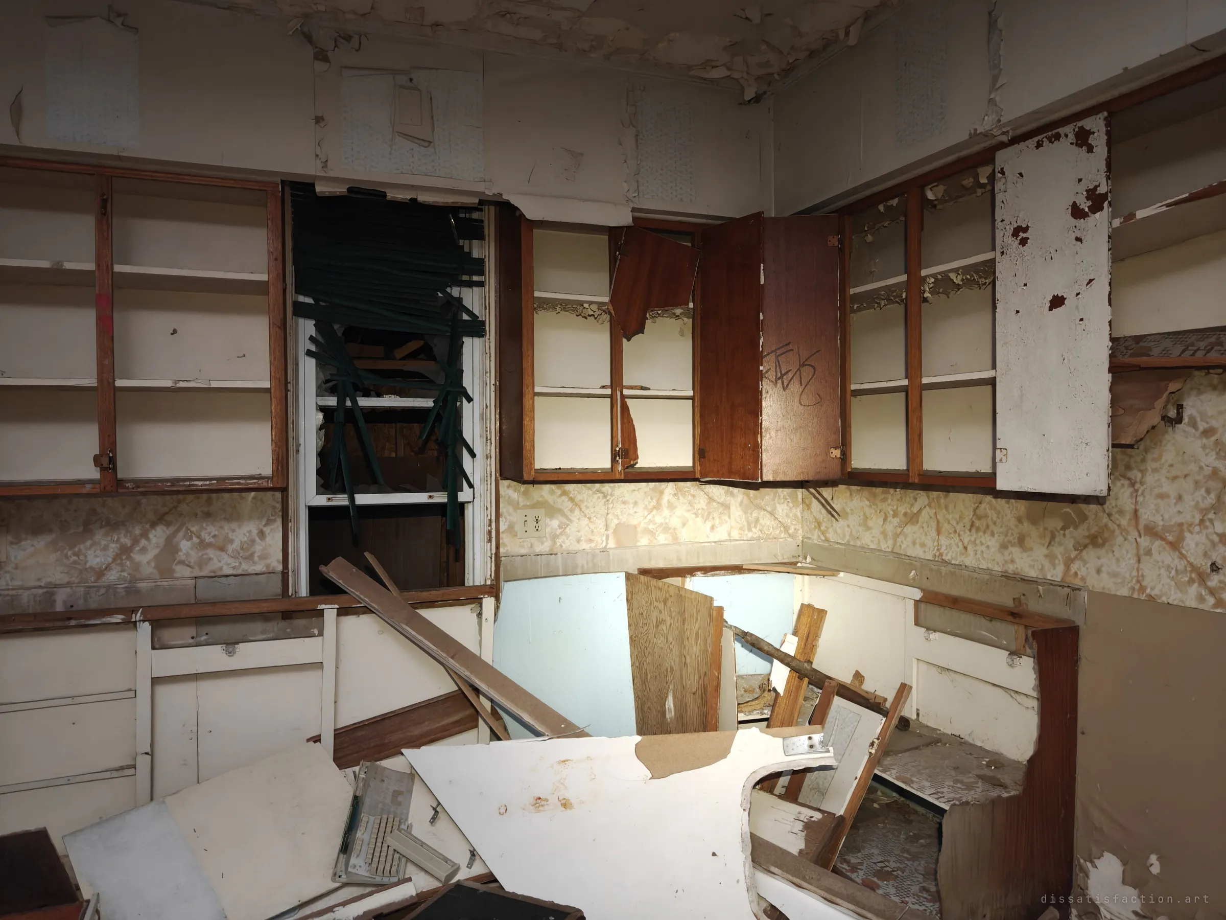 A gutted kitchen with open cabinets, peeling wallpaper and a boarded window, the counters torn out and piled with debris. The bones of a family kitchen left to rot.
