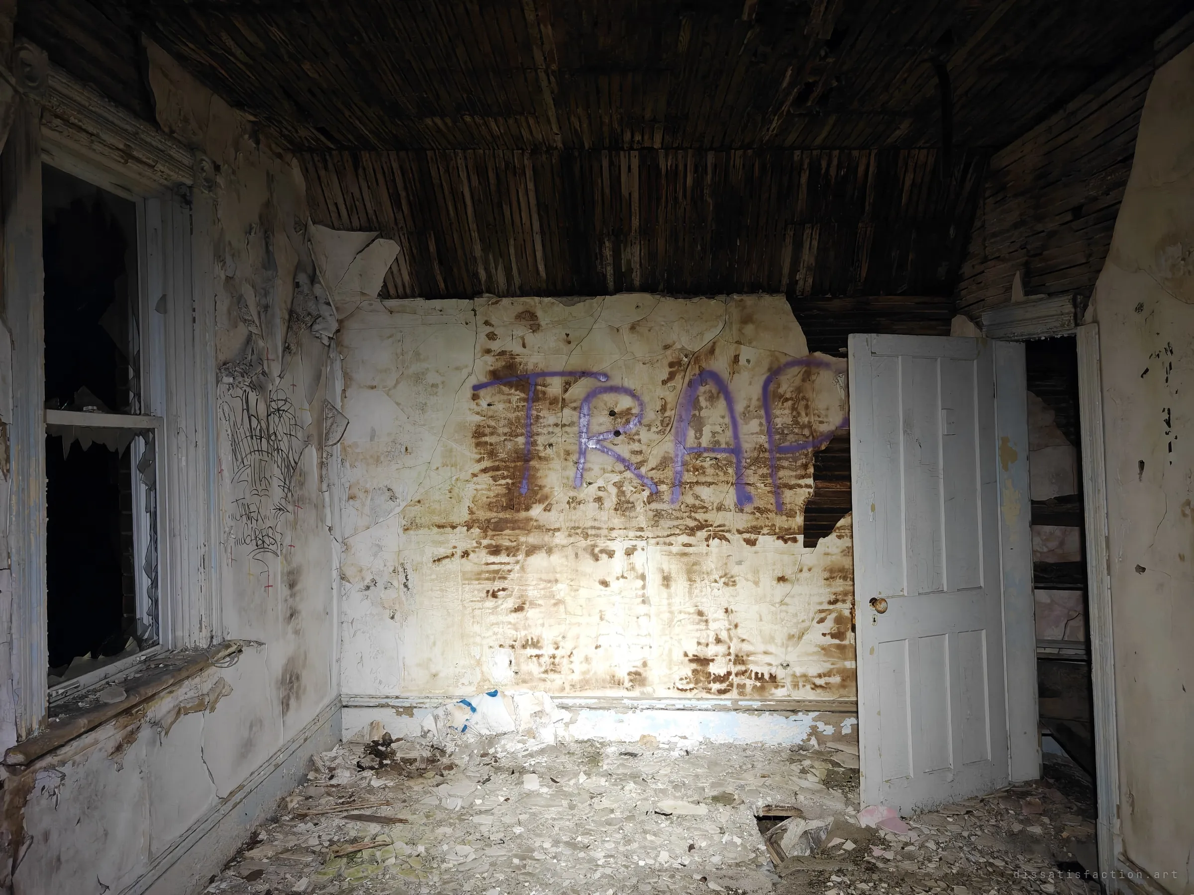 A plaster-walled room with a strip-lath ceiling, a broken window and a large purple spray-painted word across the stained back wall. Water damage and vandalism have taken over the old bedroom.