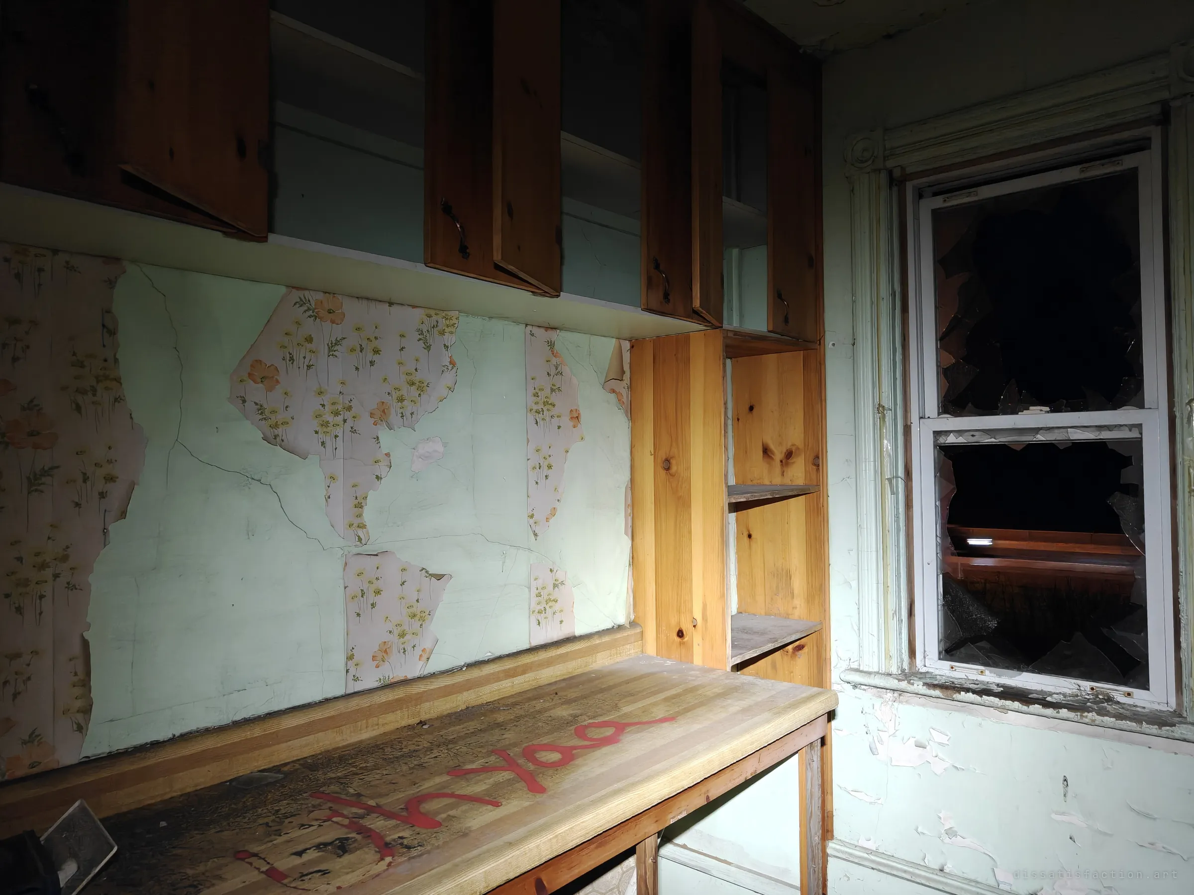 A pine countertop and cabinets under half-stripped floral wallpaper, beside a shattered sash window. Small domestic details survive among the damage.