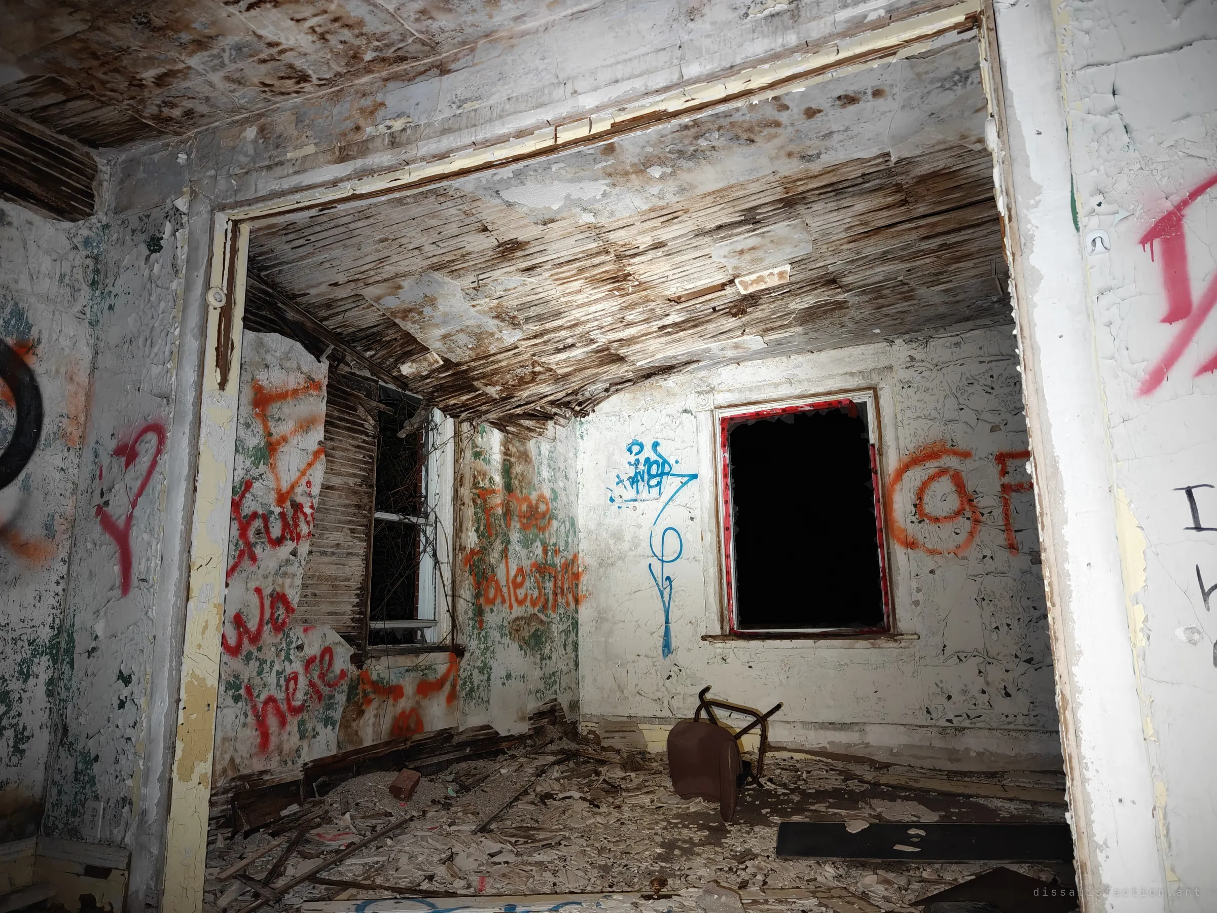 A doorway opens onto a graffiti-covered room with a sagging lath ceiling, a gaping window and an overturned chair. Layers of spray paint show how many visitors came before.