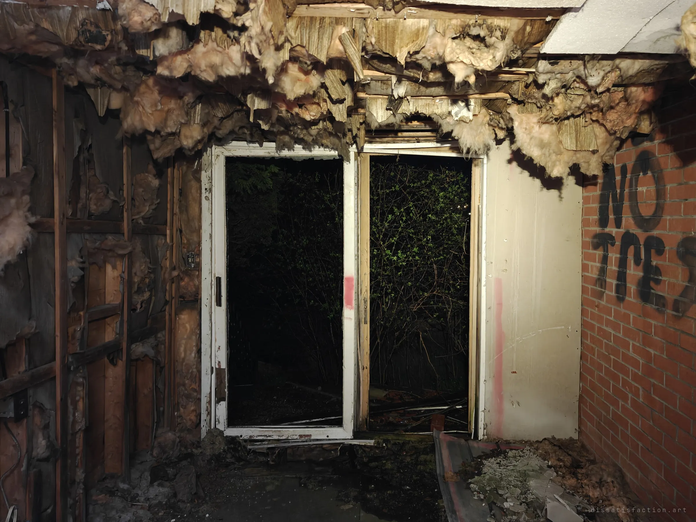 Sliding patio doors open onto dark brush, framed by torn insulation hanging from the ceiling and a brick wall sprayed with a no-trespassing warning. The back of the house is coming apart.