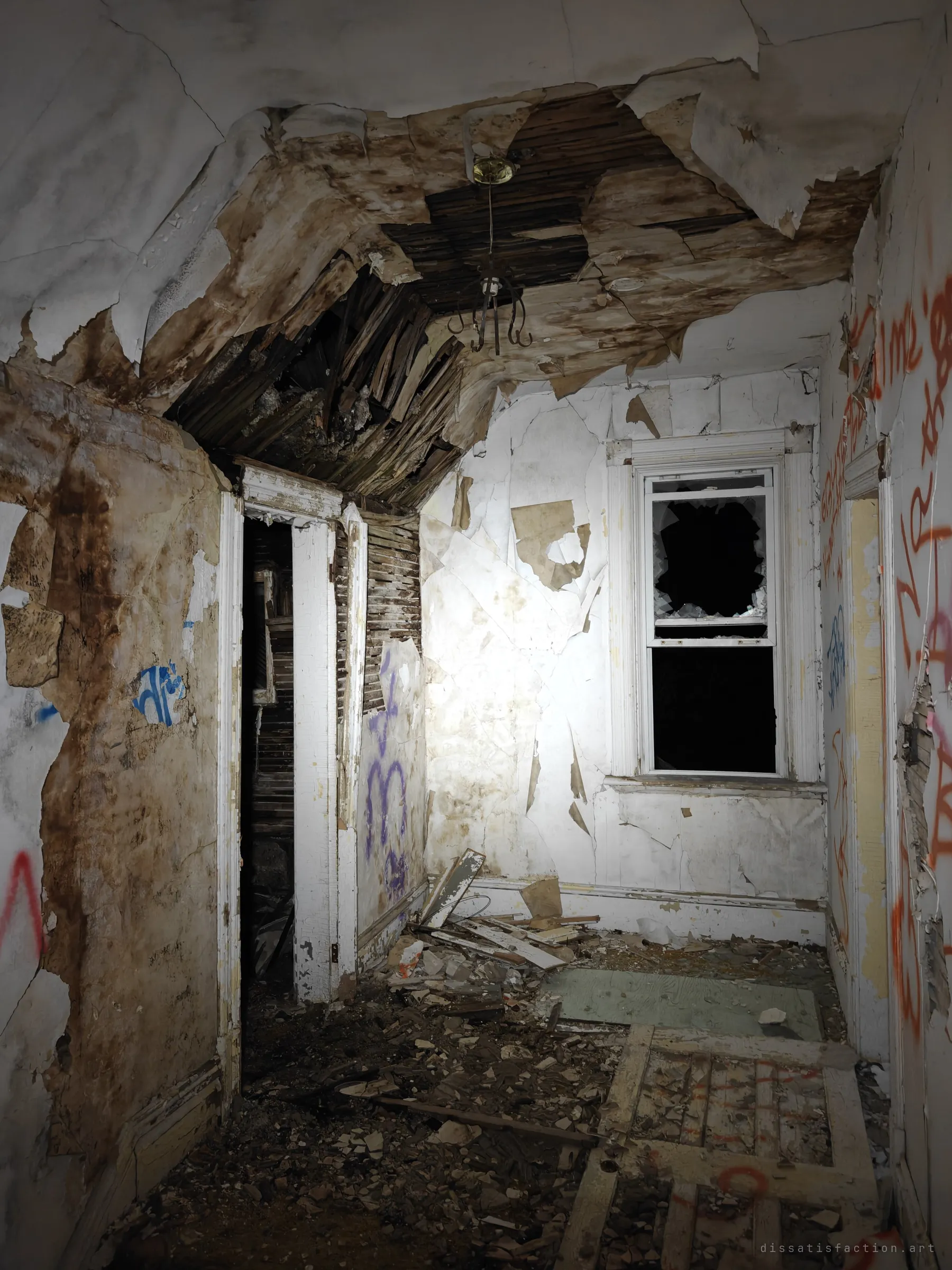 A narrow upstairs room with a collapsing ceiling, a bare light fixture and a smashed window, graffiti running down the walls. The roof is failing into the rooms below.