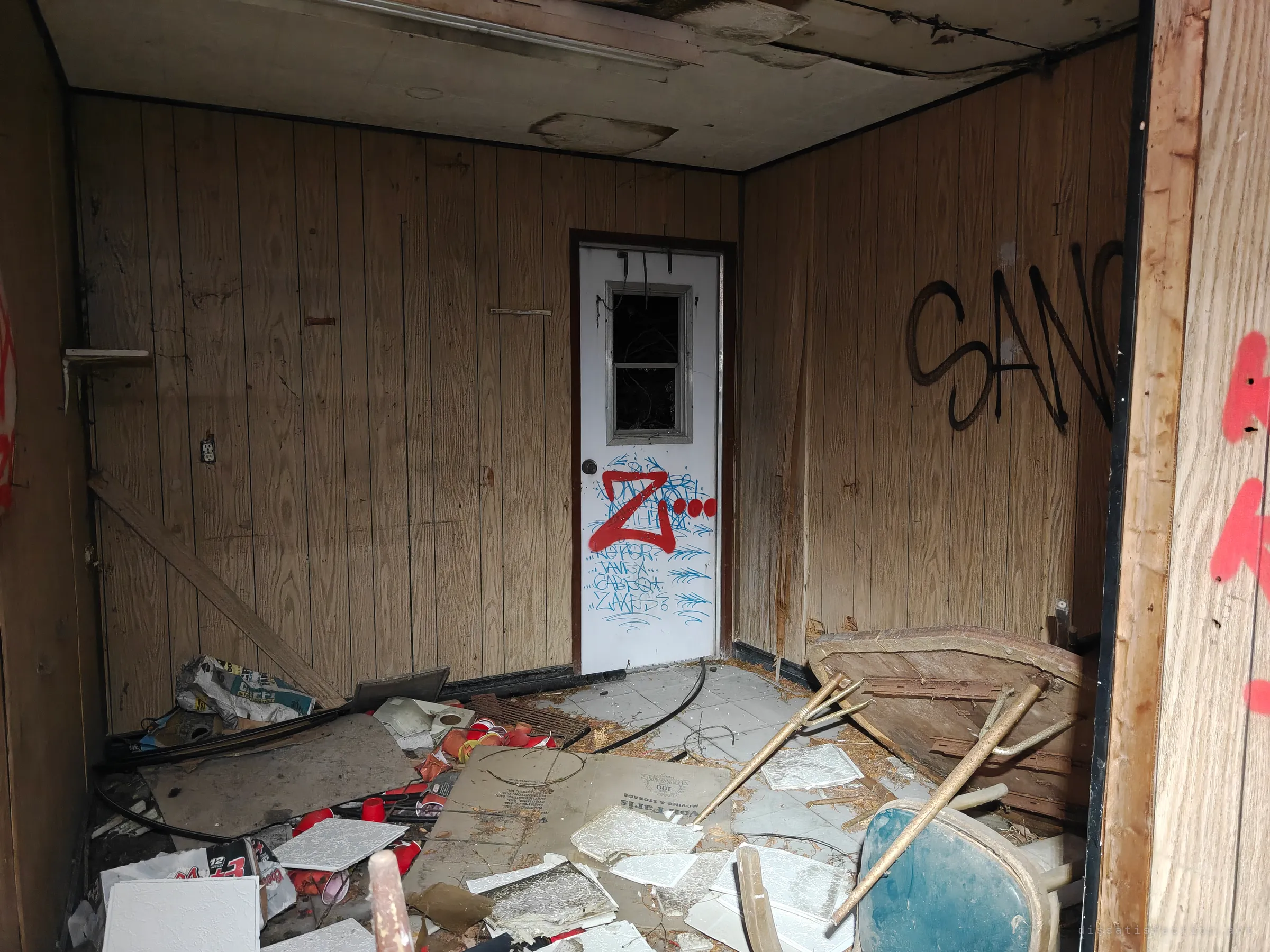 A wood-panelled back room strewn with ceiling tiles, cans and a broken chair, facing a door covered in tags. The annex was used as a hangout long after it was left.