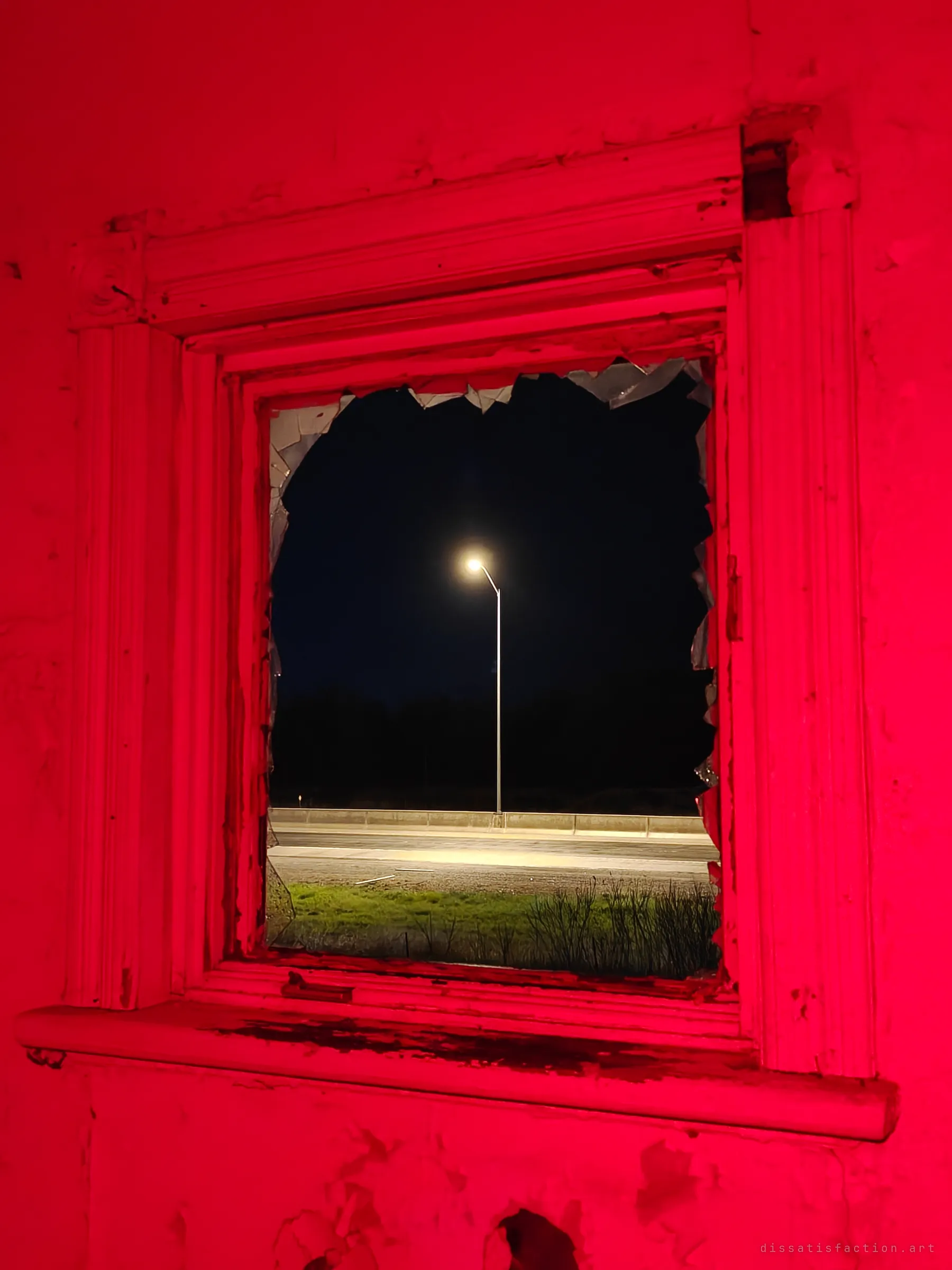 A broken window glowing red from inside frames a street lamp over an empty highway at night. The traffic outside is only a few metres from the abandoned rooms.