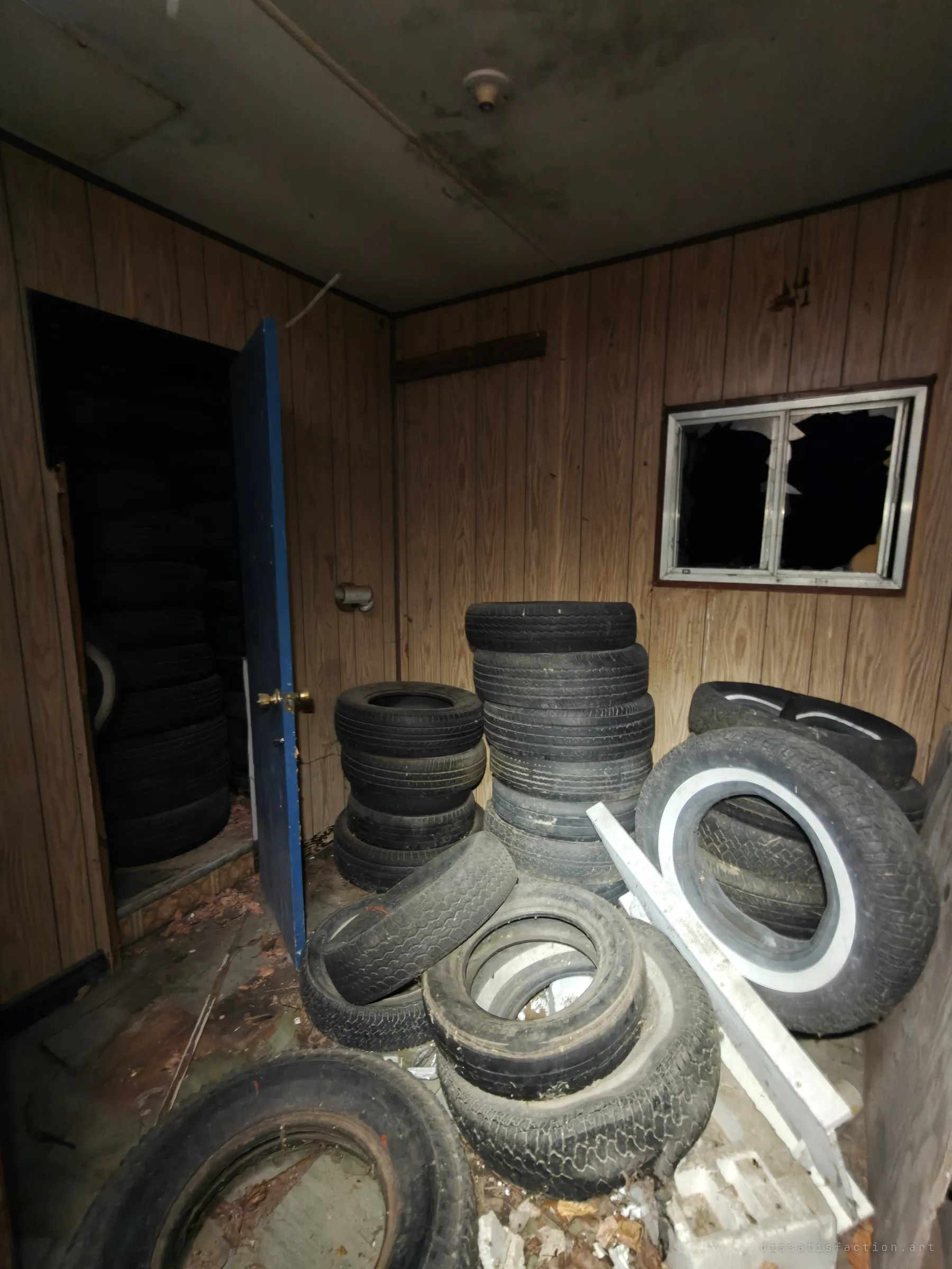 A panelled room stacked with old car tyres, some whitewalls, beside a blue door into a second room full of more. A side business seems to have ended up as storage.