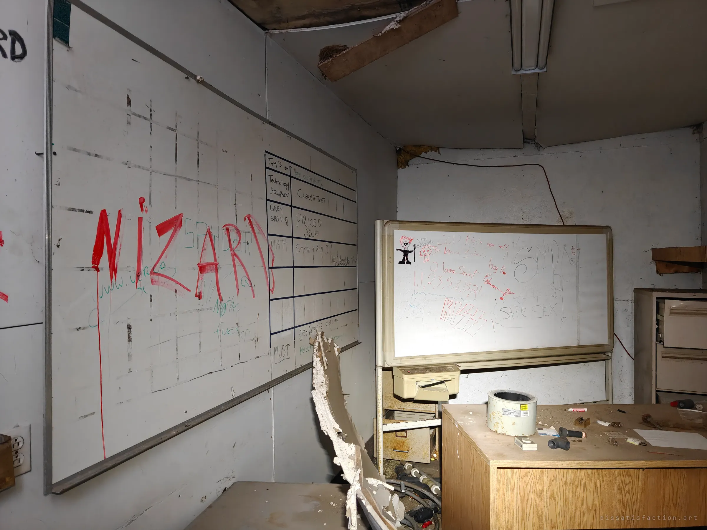 An office corner with a gridded task board and a rolling whiteboard covered in scrawled messages, a desk and filing cabinet below. Traces of a small business left behind in the annex.