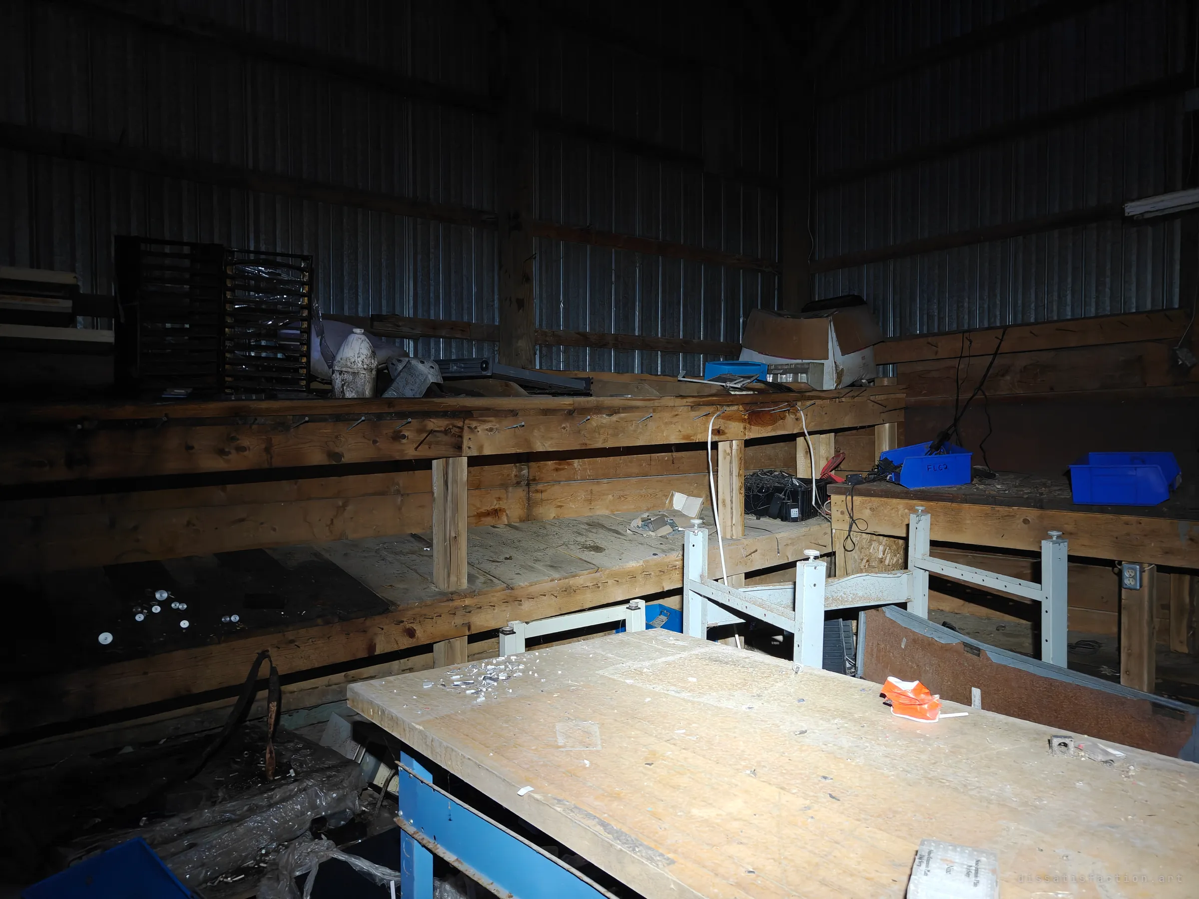 A dark workshop corner inside a tin-walled shed, lit by flashlight: long plank workbenches, blue parts bins and stray hardware left where the work stopped.