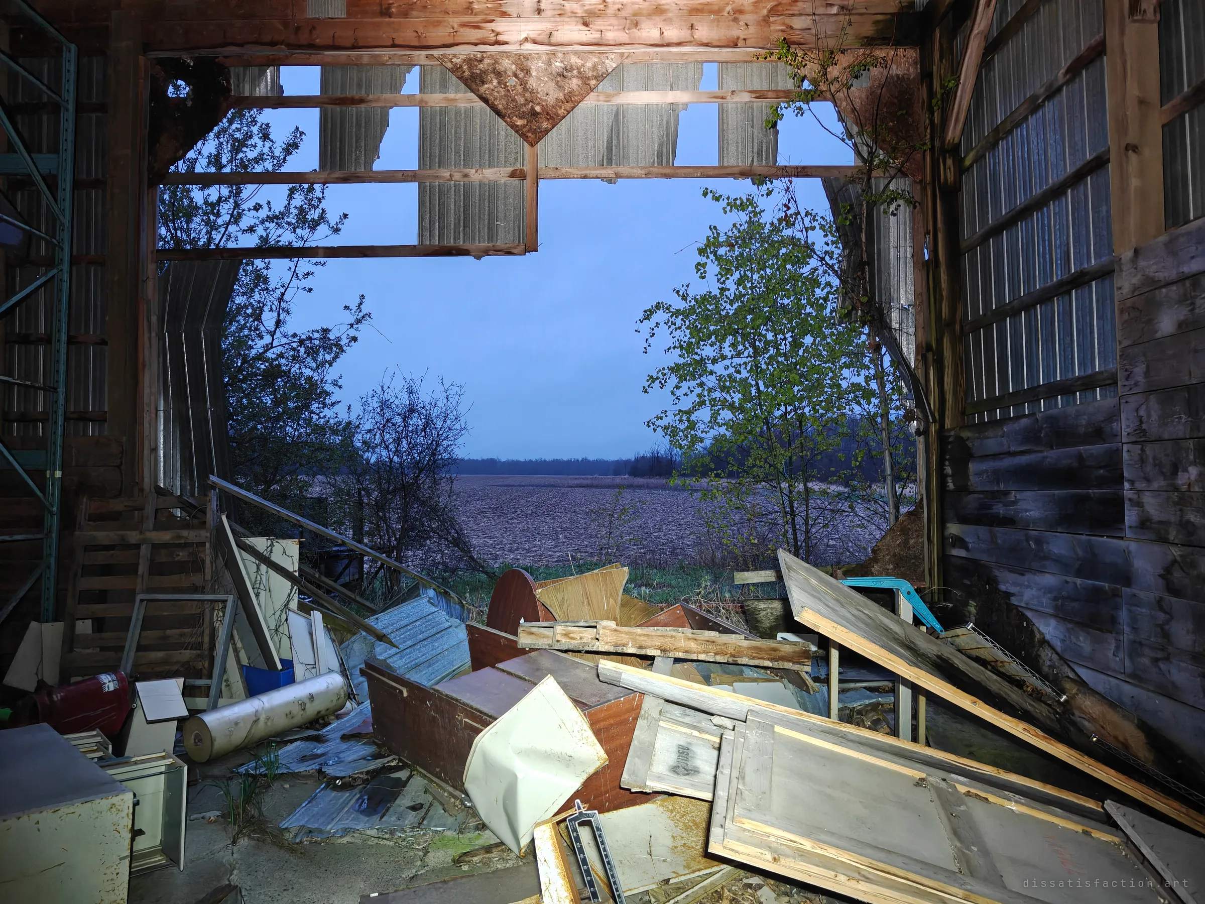 A barn wall has fallen away, framing an empty ploughed field at dusk beyond a heap of broken furniture and sheet metal. The building is slowly opening itself to the outside.