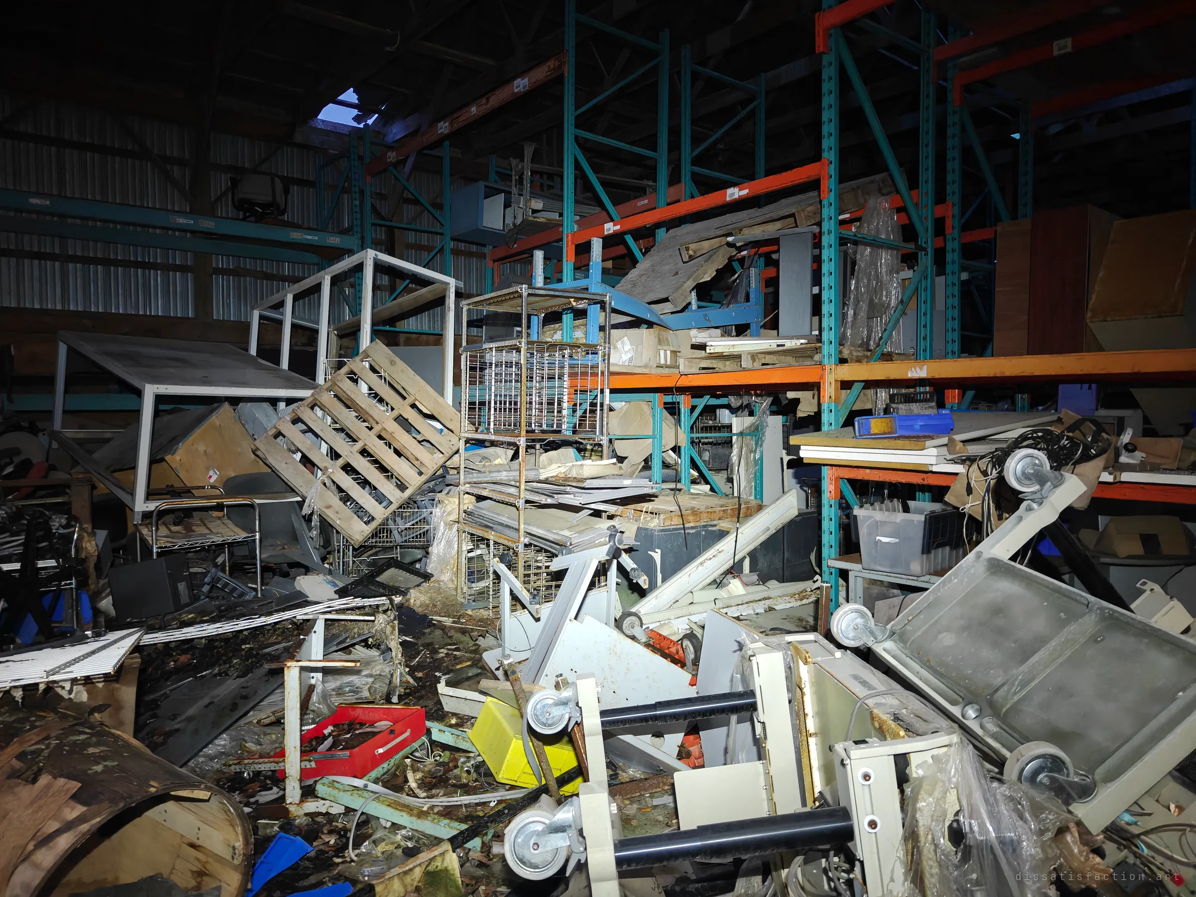 Teal and orange pallet racking towers over a tangled pile of carts, wire shelving and crates, with a hole in the roof letting in the evening sky.