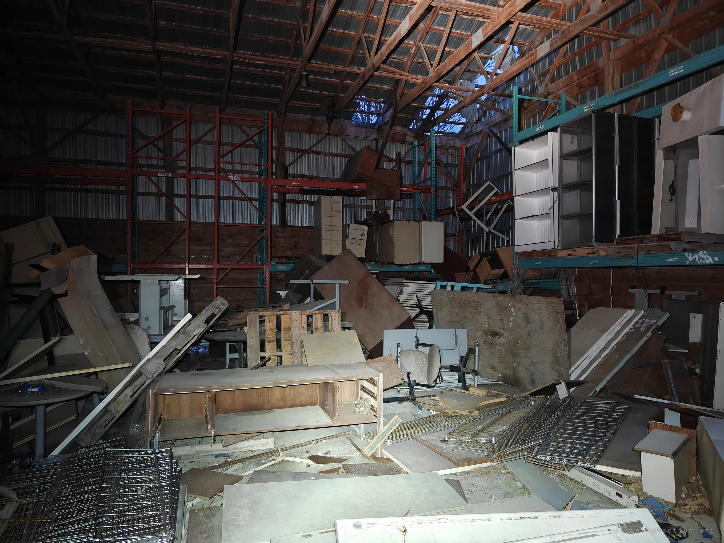 A cavernous storage barn under red timber trusses, its floor buried in desks, filing cabinets, plywood and wire racks; daylight leaks through a torn section of roof.