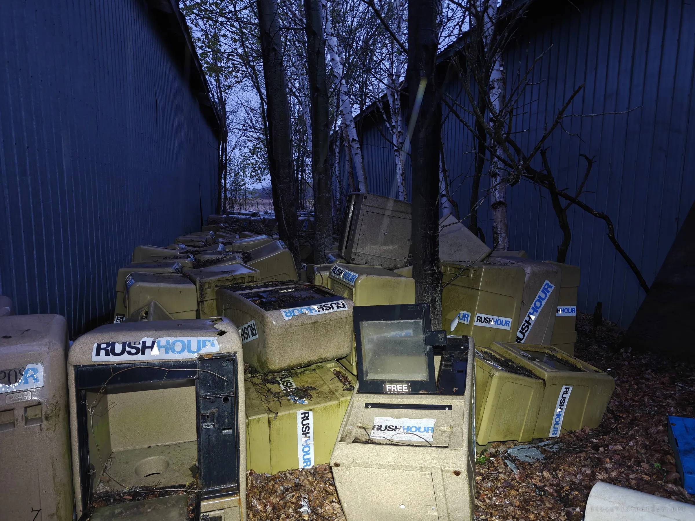 A narrow gap between two corrugated sheds is packed with abandoned street newspaper boxes, stacked and toppled among birch trees. A small graveyard of a paper nobody prints anymore.
