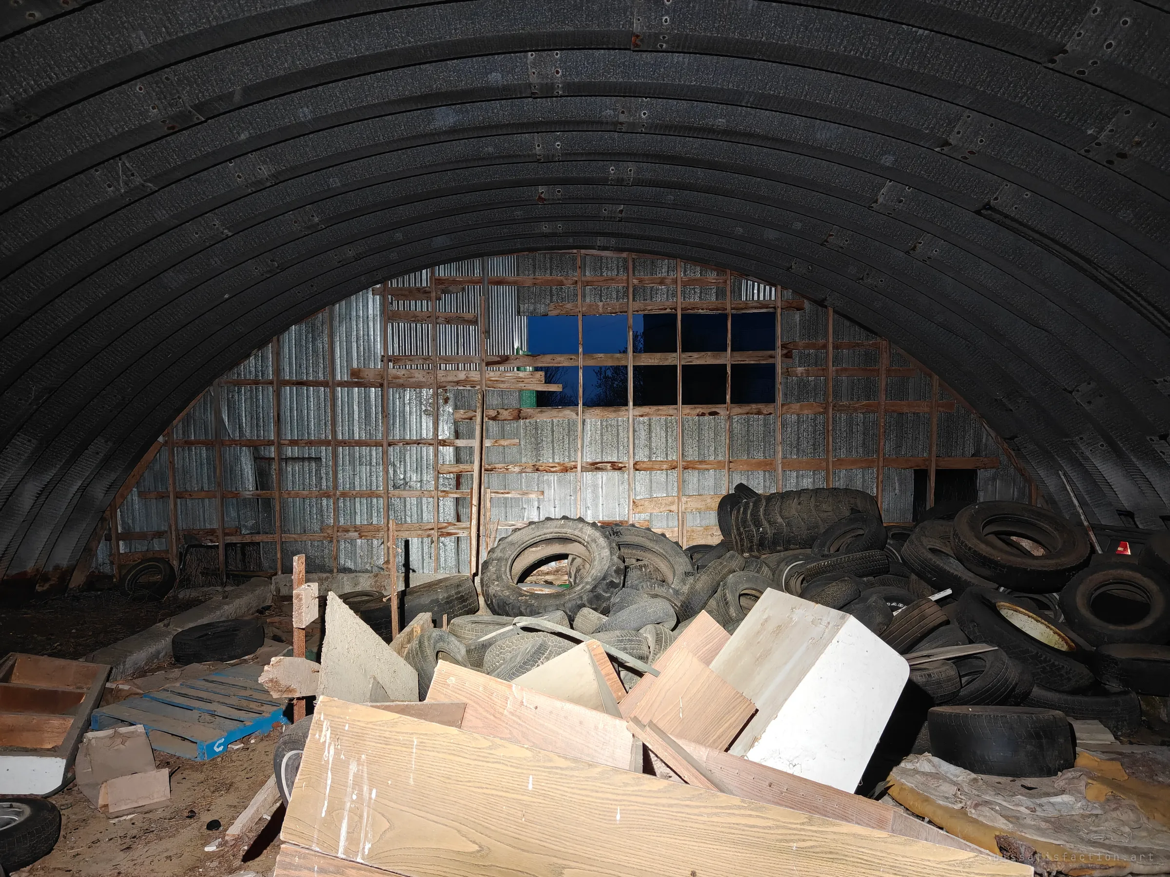 Inside a ribbed steel Quonset hut, an arched ceiling curves over a mound of old tyres and loose lumber, with a stripped end wall open to the night.