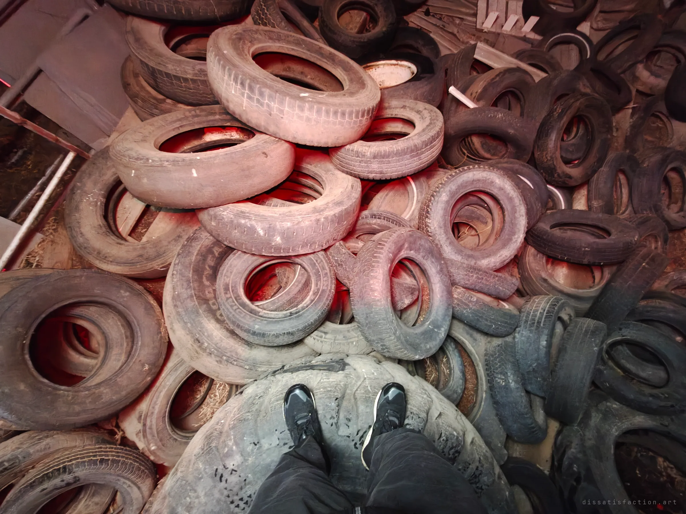 Looking straight down at the photographer's shoes standing on a huge tractor tyre in a sea of discarded tyres, washed in red light.