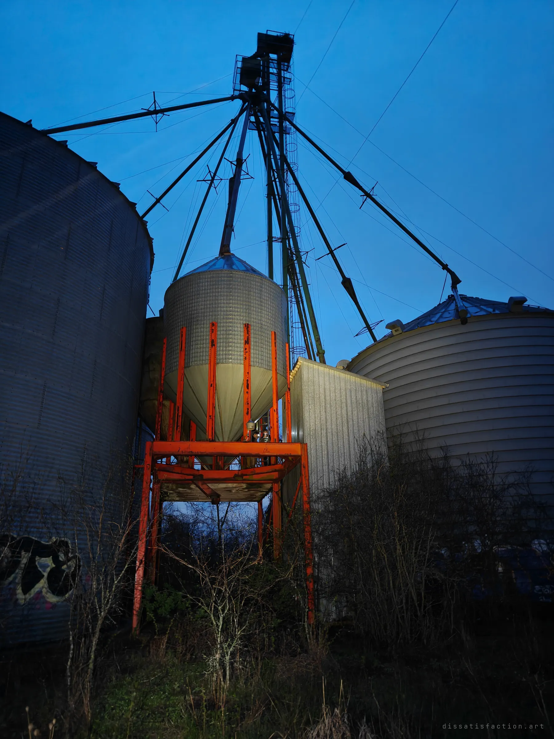 Looking up at a hopper bin on a rust-orange steel stand, wedged between two larger grain bins beneath a web of elevator spouts and a deep blue sky.