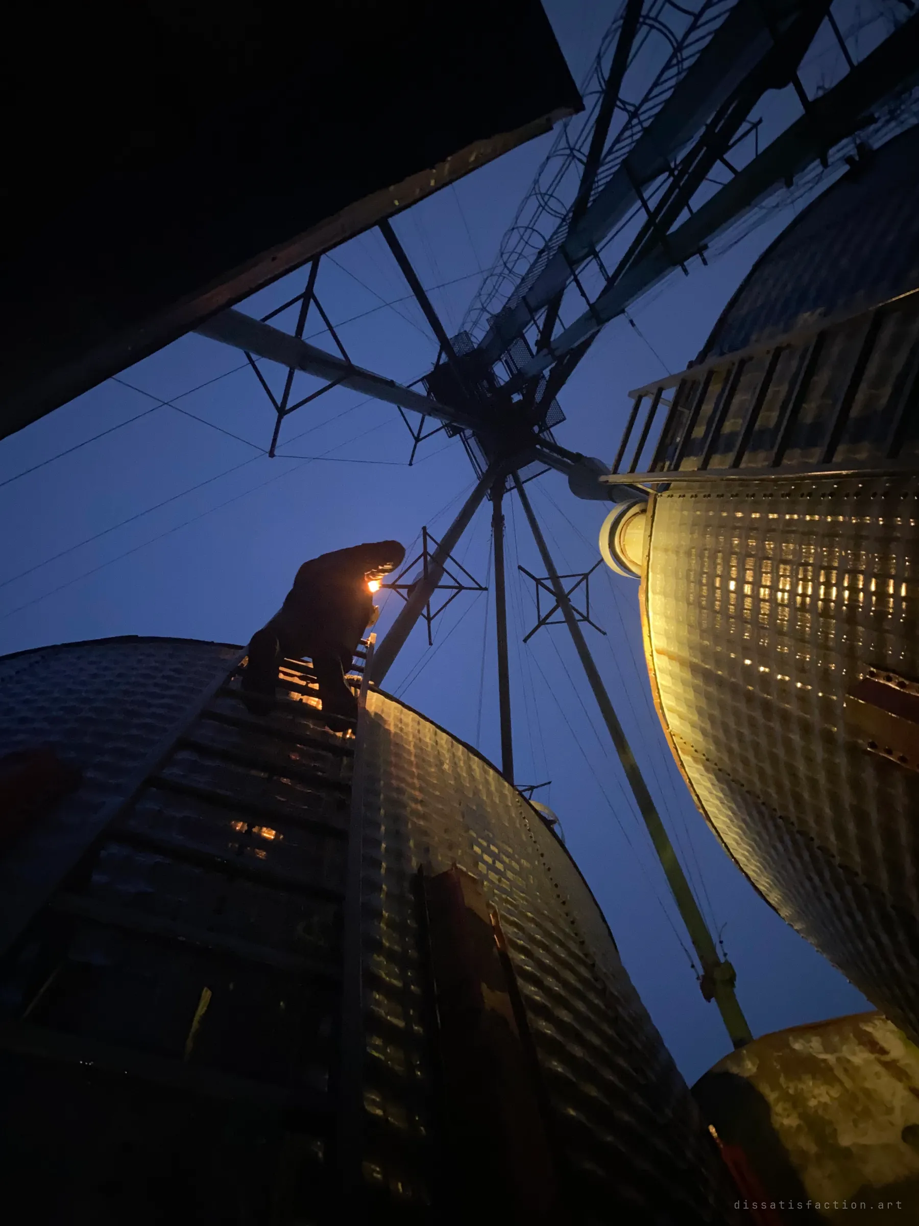 From below, an explorer climbs a ladder up the side of a lamp-lit grain bin, silhouetted against the night sky and the radiating spouts of the elevator.