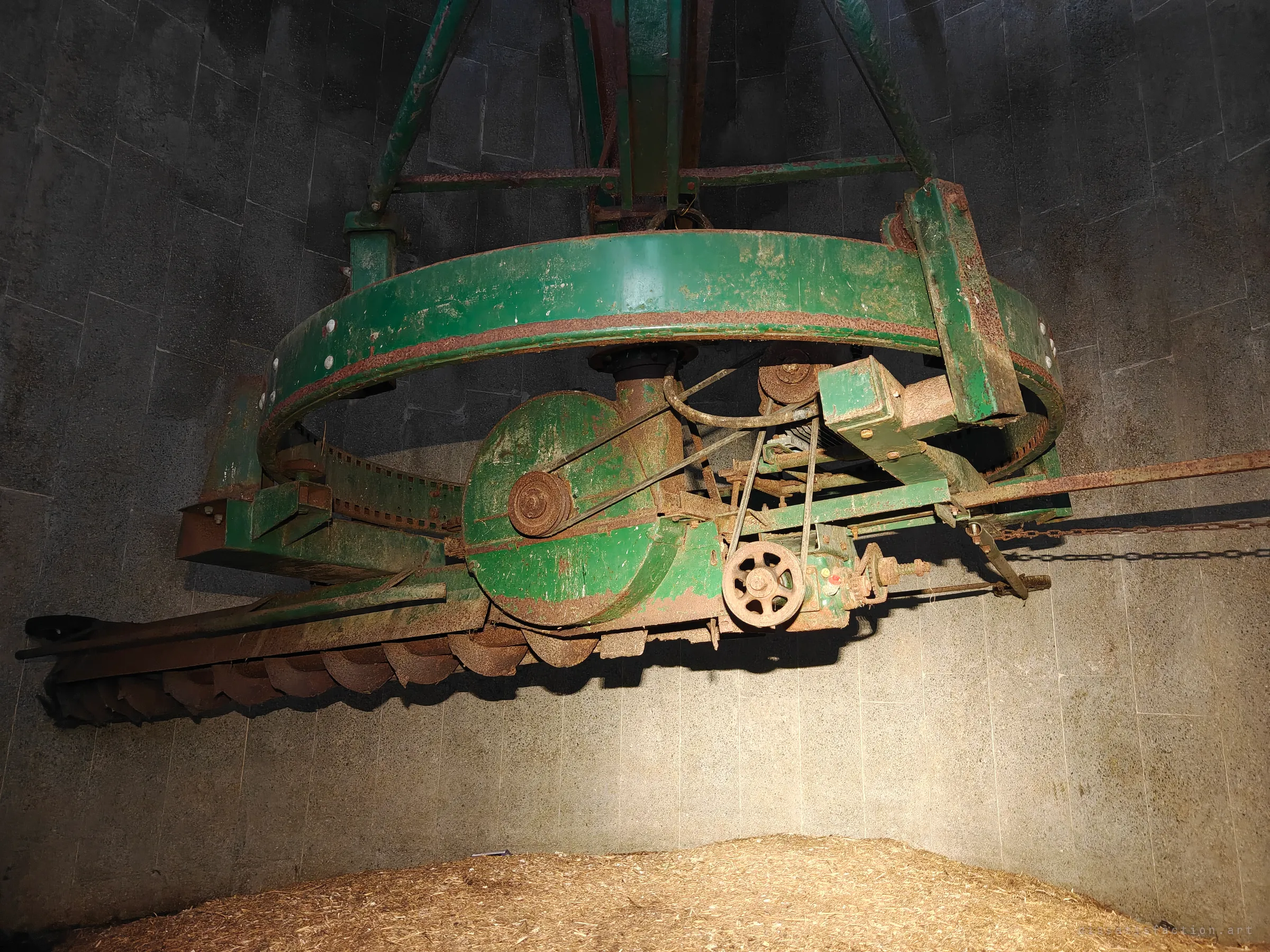 Inside a concrete silo, a rusted green unloader hangs over a heap of old feed, its gears, belts and auger frozen mid-turn.