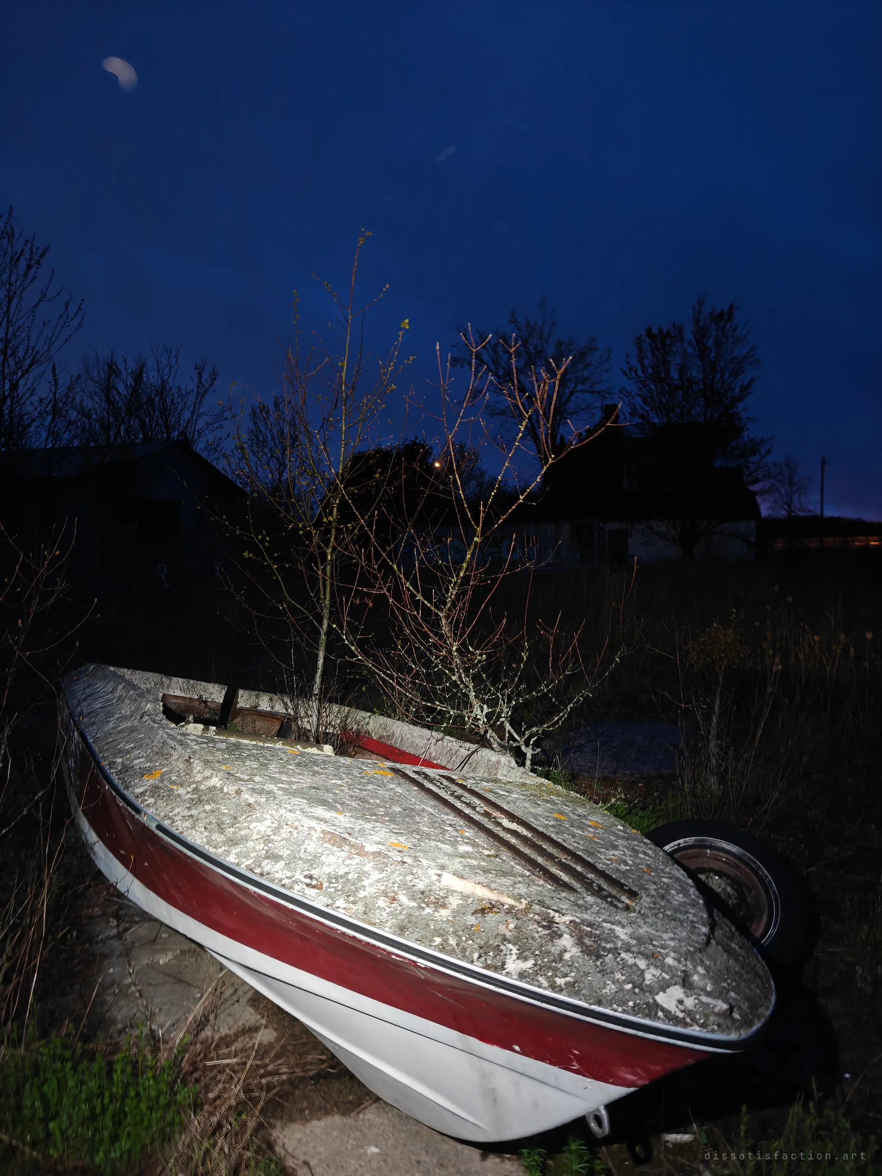 An abandoned speedboat on a trailer sits lichen-covered in overgrown grass, with a young tree growing out of its cockpit. Dusk sky and dark farm buildings sit behind it.