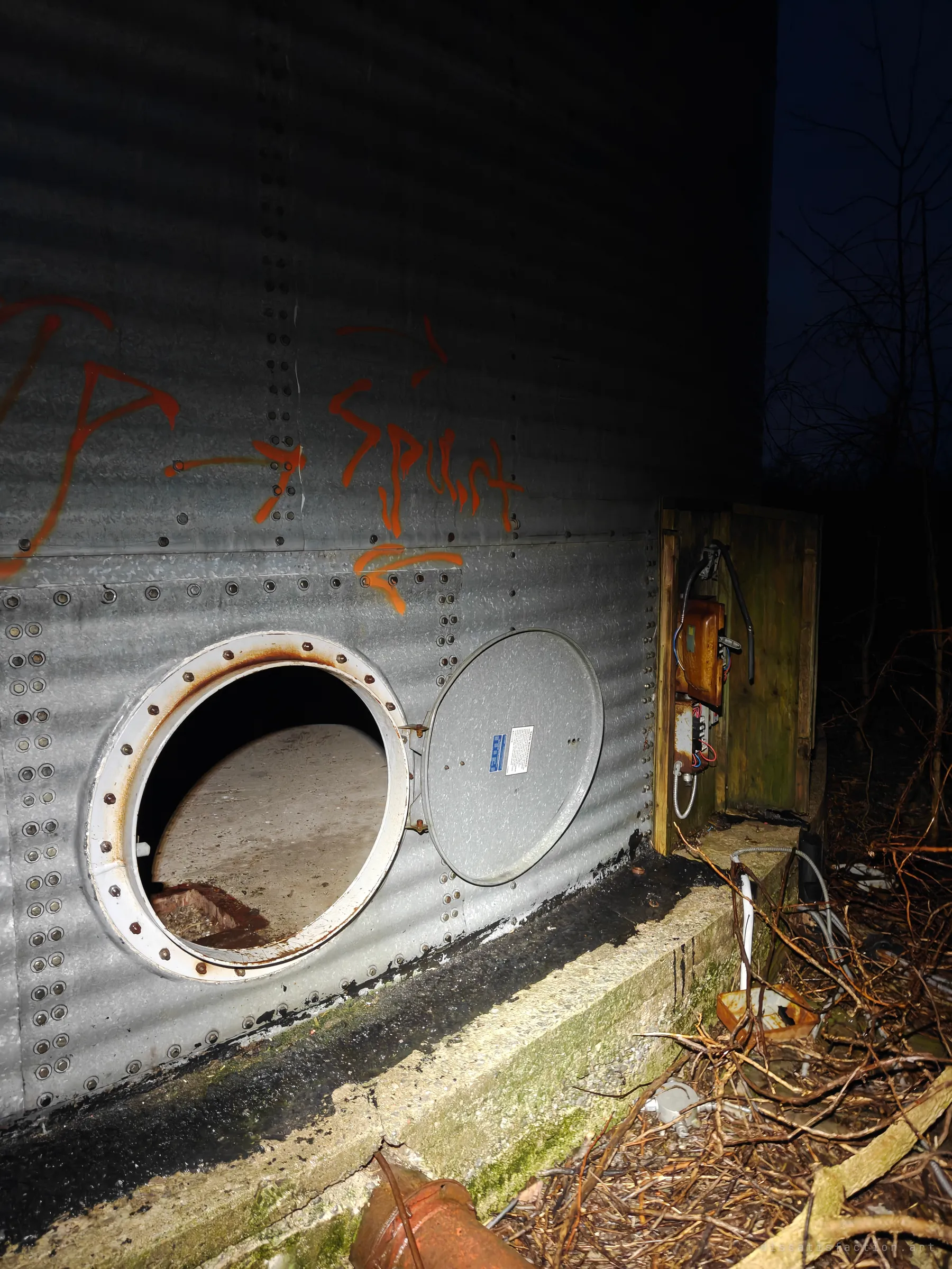 A round hatch stands open on the base of a corrugated steel grain bin, beside a rusted electrical box in a wooden enclosure. Graffiti arrows and a tag mark the entry point.