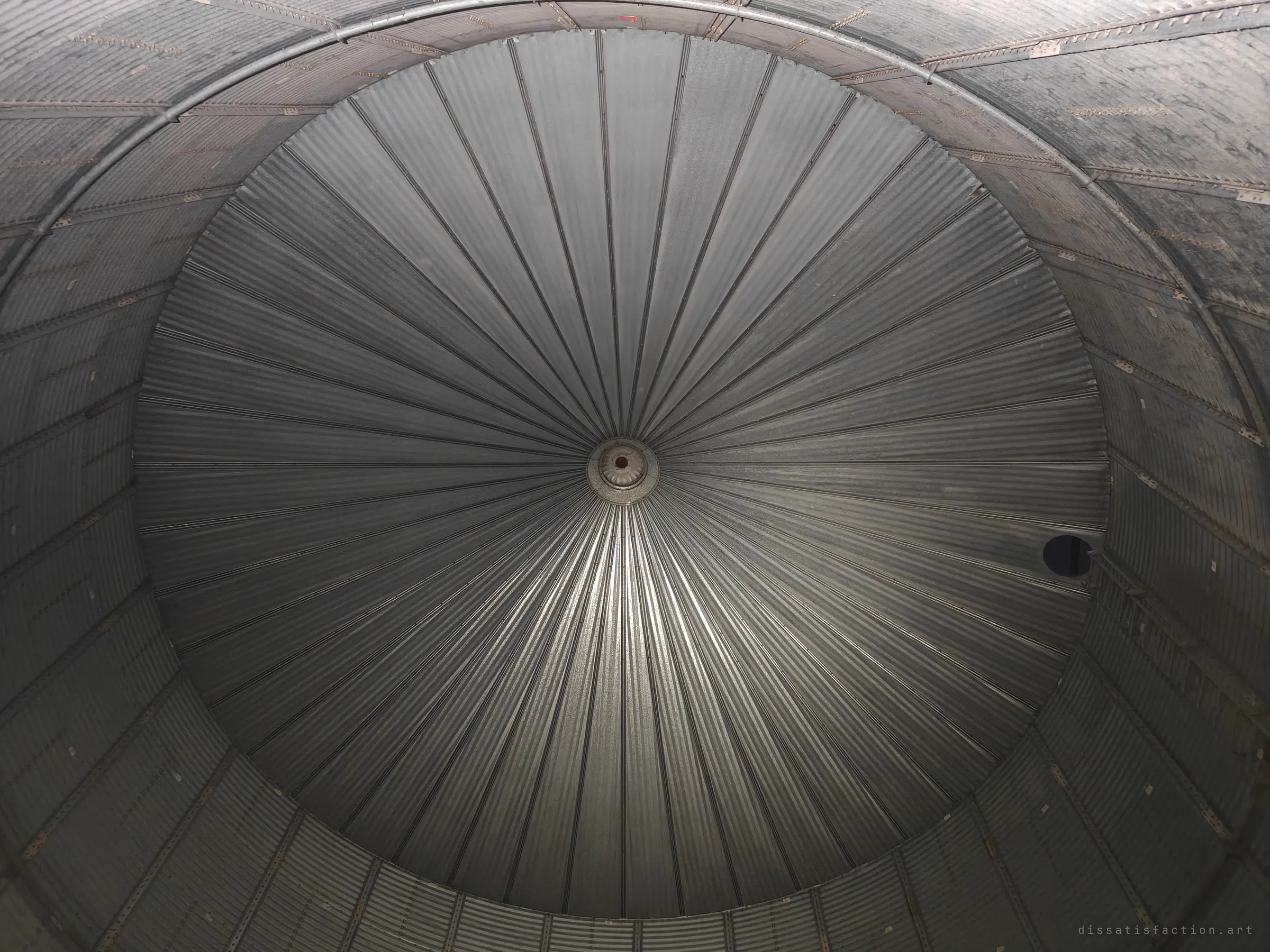 From inside the empty grain bin, the corrugated roof radiates from a central vent like a giant aperture. The symmetry turns a farm structure into something cathedral-like.