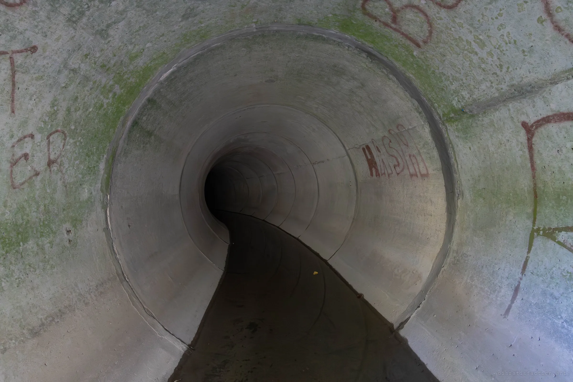A round concrete drain mouth with green algae and red spray-painted graffiti on its face, its pipe curving off to the left into darkness. The S-bend in the dark floor channel pulls the eye into the tunnel.
