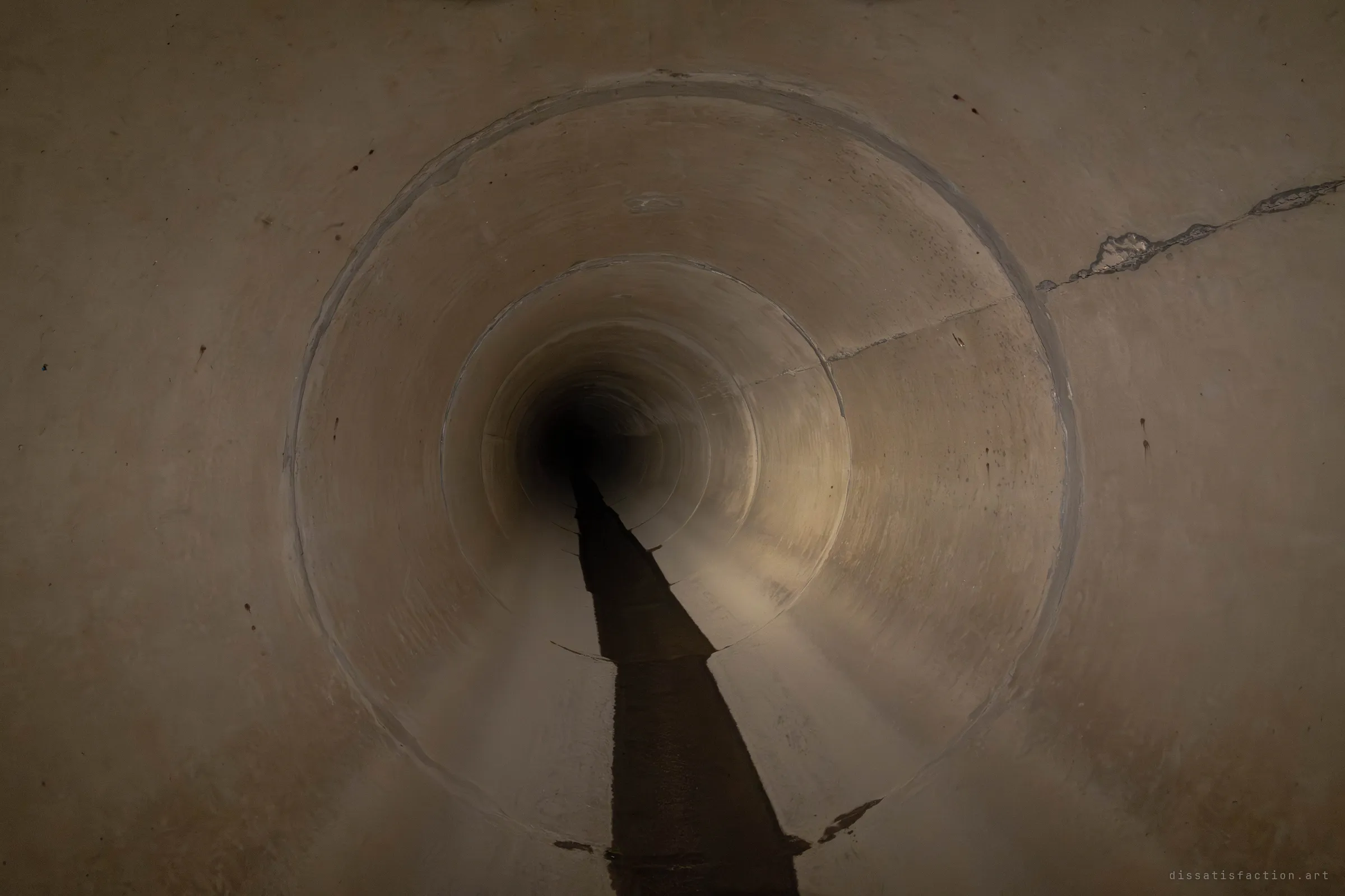 A smooth tan concrete pipe telescopes down into a smaller tunnel, with a dark wet trickle running straight down the floor. A low mist hangs in the torchlight over the invert.