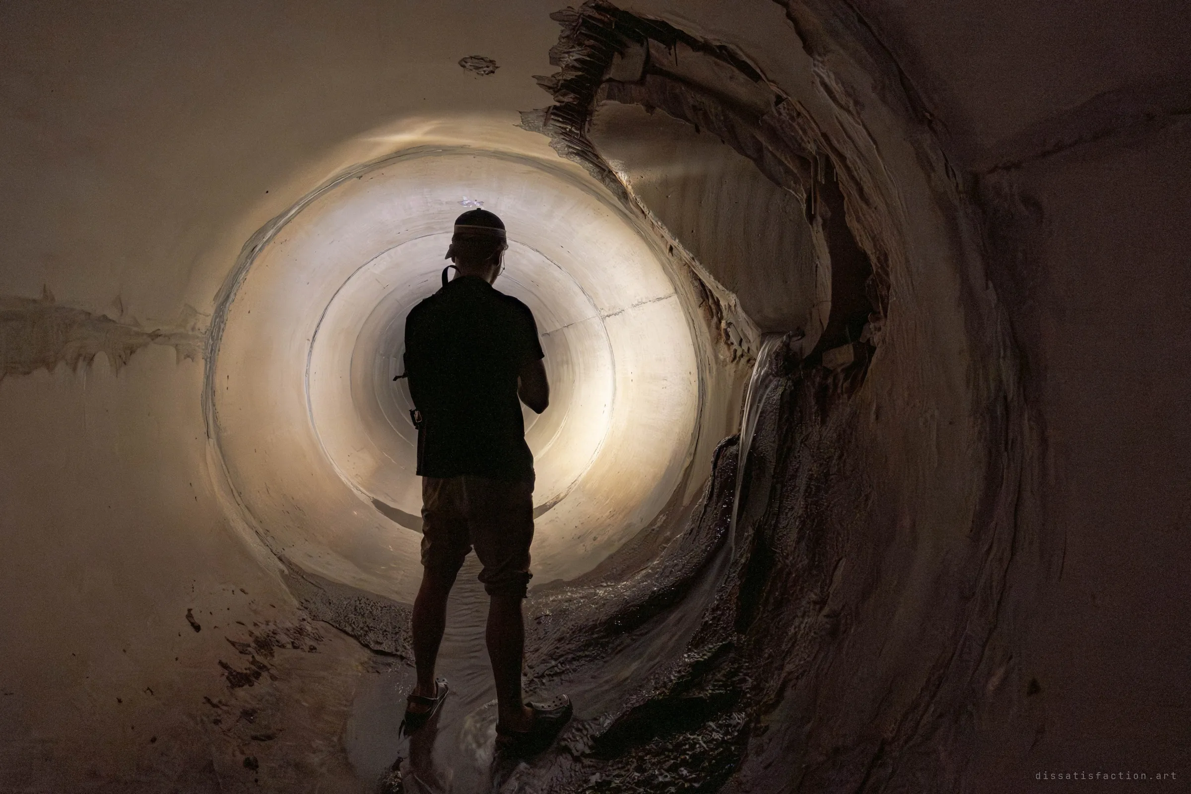A silhouetted explorer in a headlamp and shorts stands in a round concrete storm drain, facing a glowing tunnel ahead. To the right, the lining has peeled away in layers and water trickles down the torn wall.