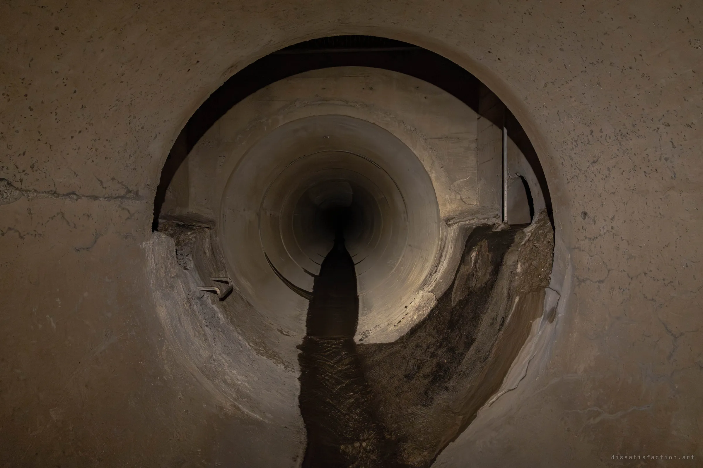 Seen through a larger circular opening, a lit concrete pipe runs away into the dark past a chamber with a thin vertical pole and a rusted step rung. Silt banks either side of the dark water channel show where the flow has cut its own path.