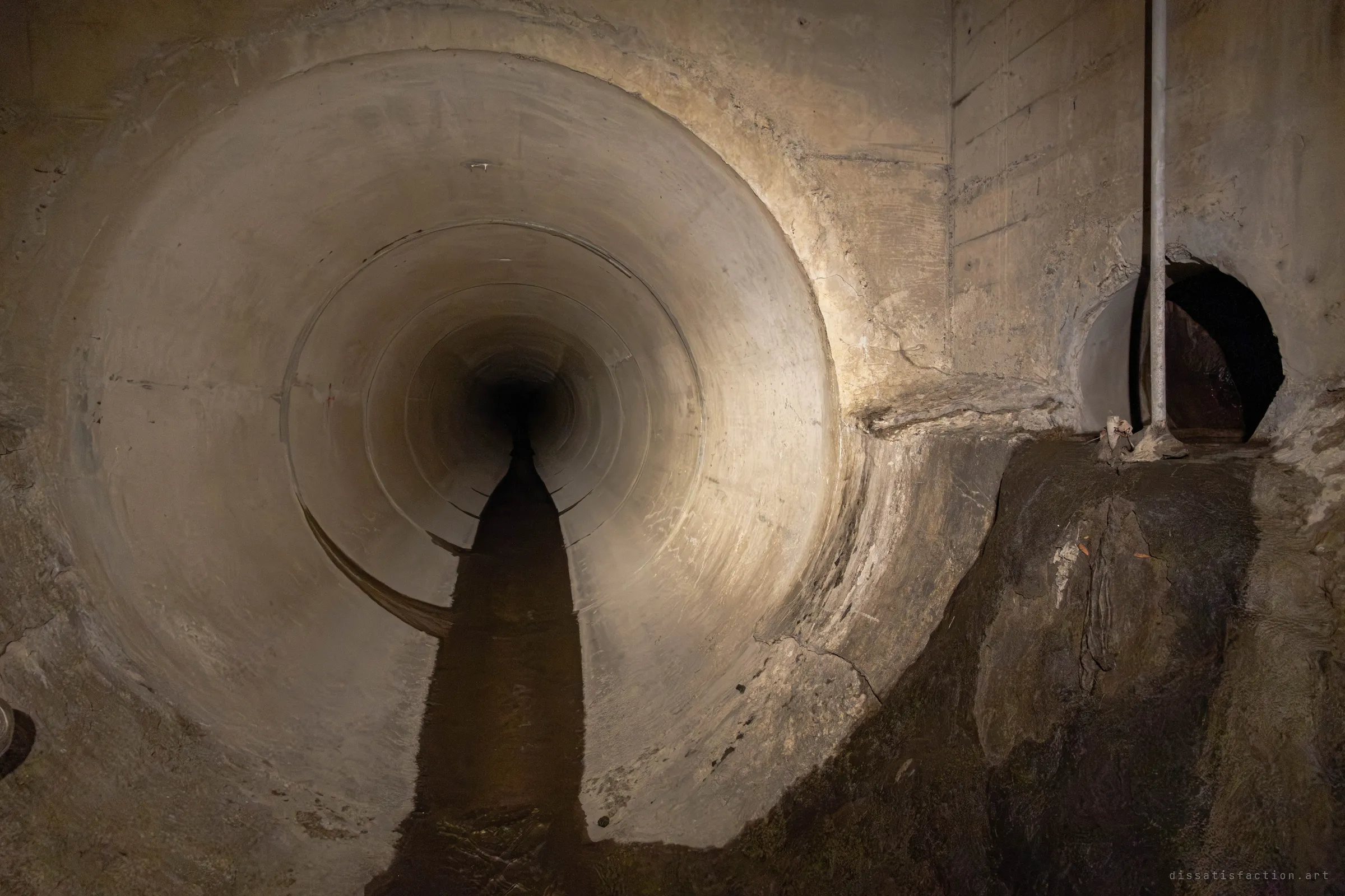A torch-lit pipe mouth glows warm against a rough board-formed concrete chamber, with a side inlet and a vertical pole to the right above a bank of dark silt. The water channel runs out of the bright pipe toward the camera.